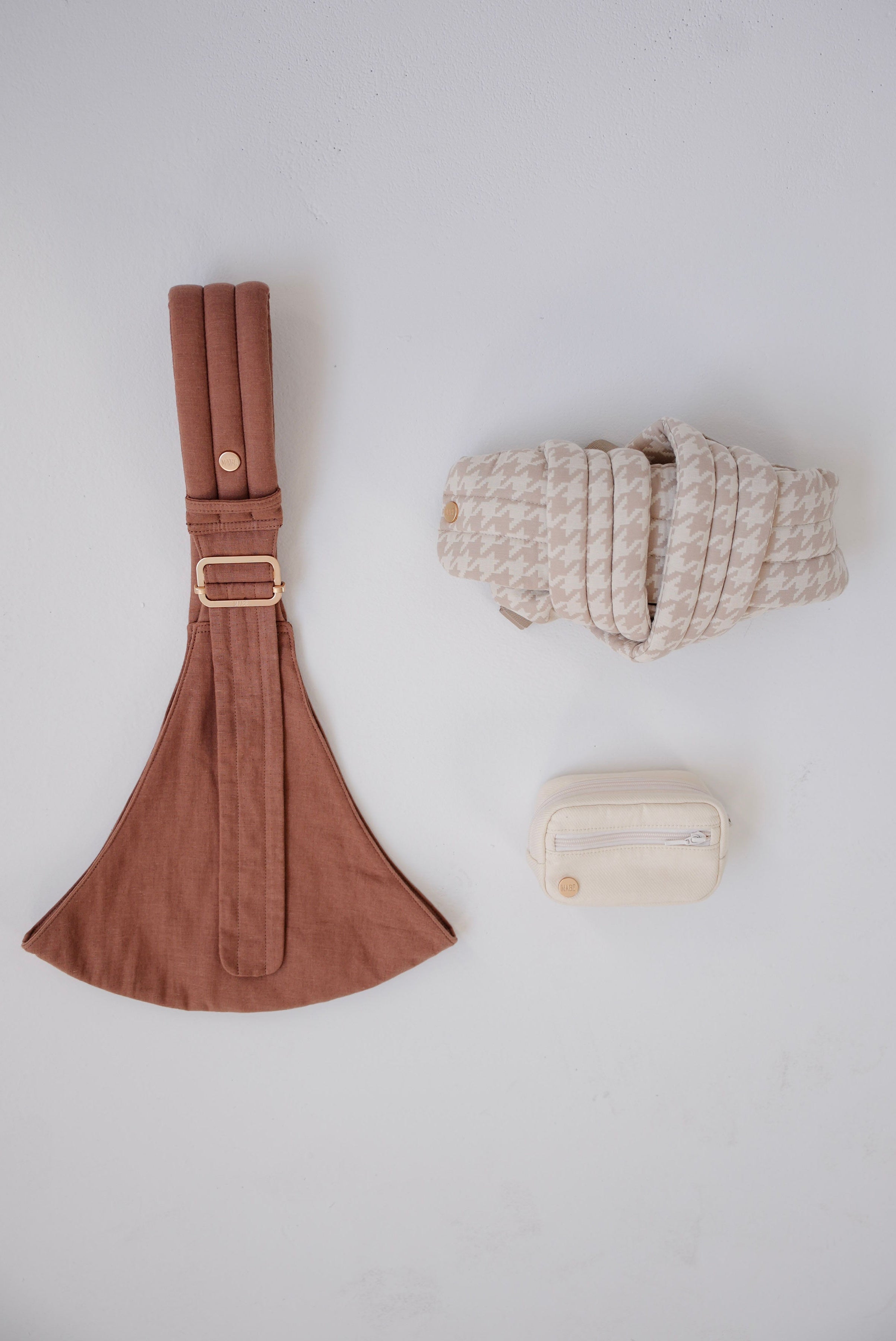 Solid mocha-colored Mabē toddler sling with a rolled-up beige houndstooth patterned Monarch buckle carrier and solid ivory accessory bag on a white background