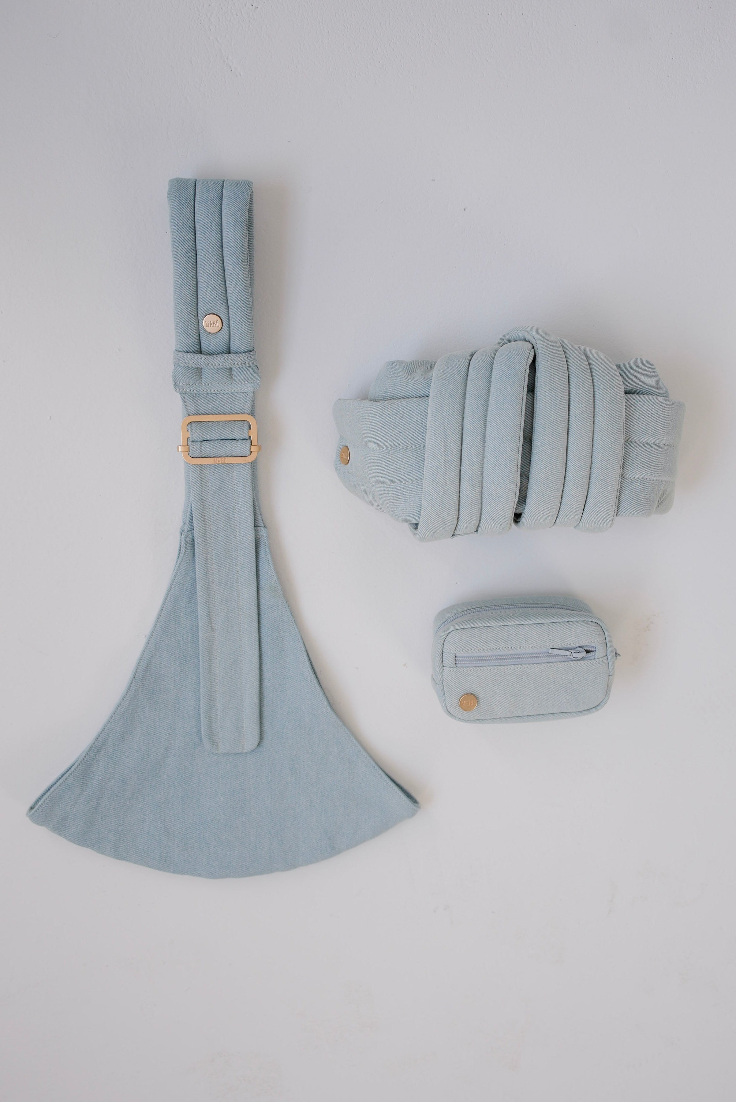 Light blue denim Mabē toddler sling with a matching rolled-up Monarch buckle carrier and accessory bag on a white background