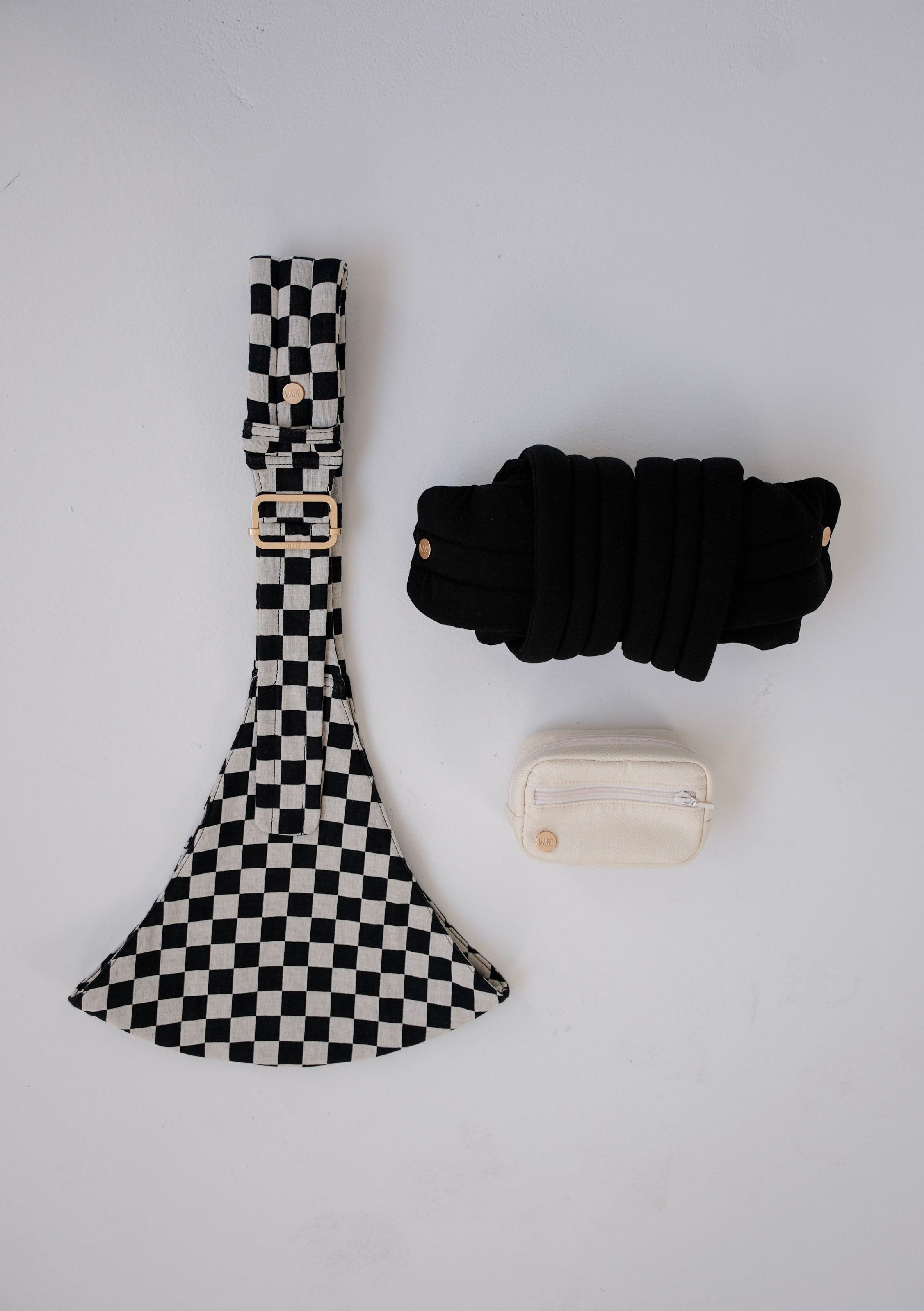 Black and white checkered Mabē toddler sling with a solid black rolled-up Monarch buckle carrier and solid ivory accessory bag on a white background