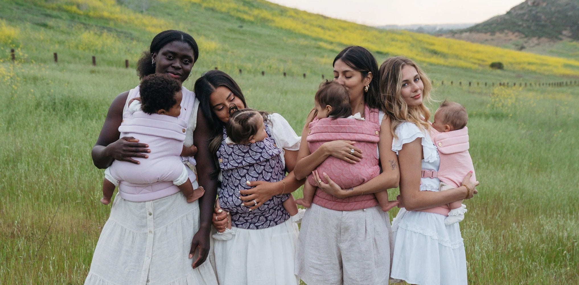 Four women in a field with wildflowers and a fence. They all wear white clothing and babies in Mabē Monarch buckle carriers in solid lilac, dark purple with butterfly wings, medium pink with butterfly wings, and solid light pink. The first woman has dark skin and black hair. The second and third have straight dark hair and tan skin. The fourth has brown and blonde hair, light skin.