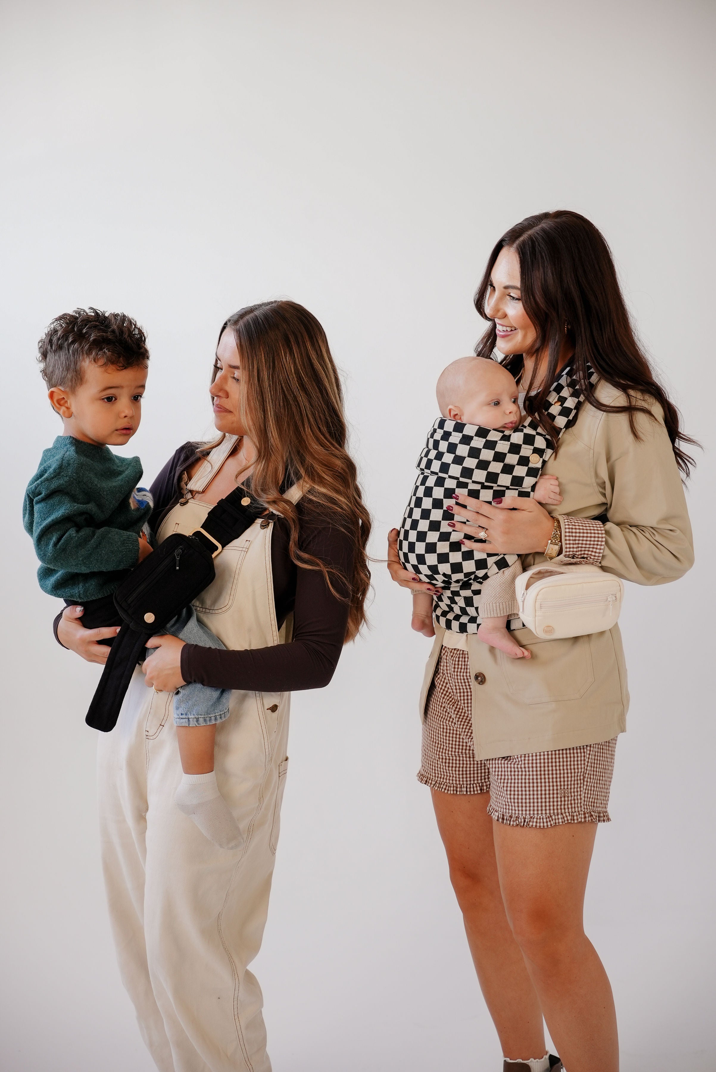 Two white women with long brown hair, one with highlights, holding children using Mabē baby carriers against a white background. The woman with highlighted hair holds a tan-skinned toddler boy in a dark green shirt in a solid black toddler sling; the other woman, who is smiling, holds a light-skinned baby in a black-and-white checkered Mabē Monarch buckle carrier.