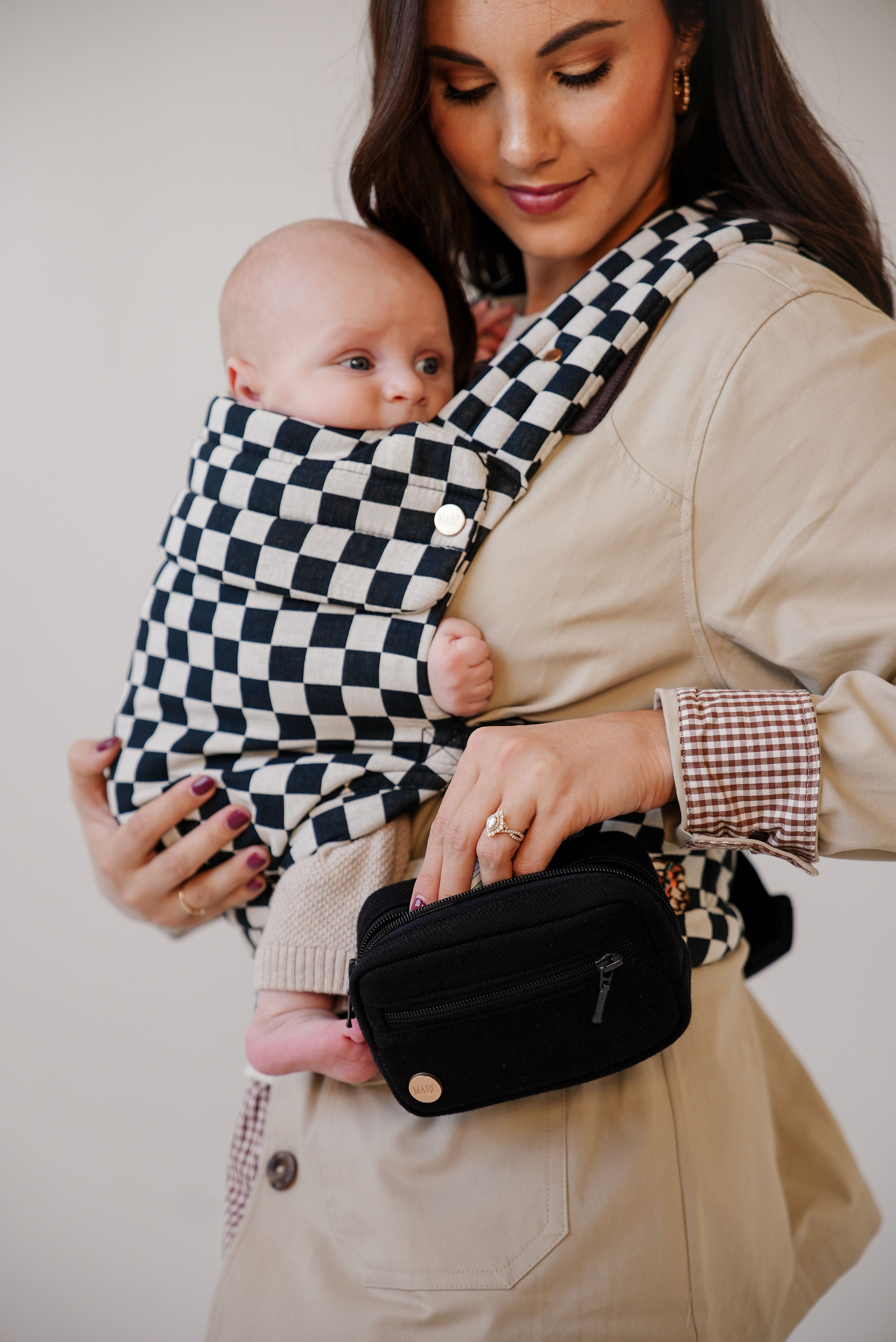 A tan-skinned woman with long brown hair holding a light-skinned baby in a black-and-white checkered Mabē Monarch buckle carrier with a black accessory bag.