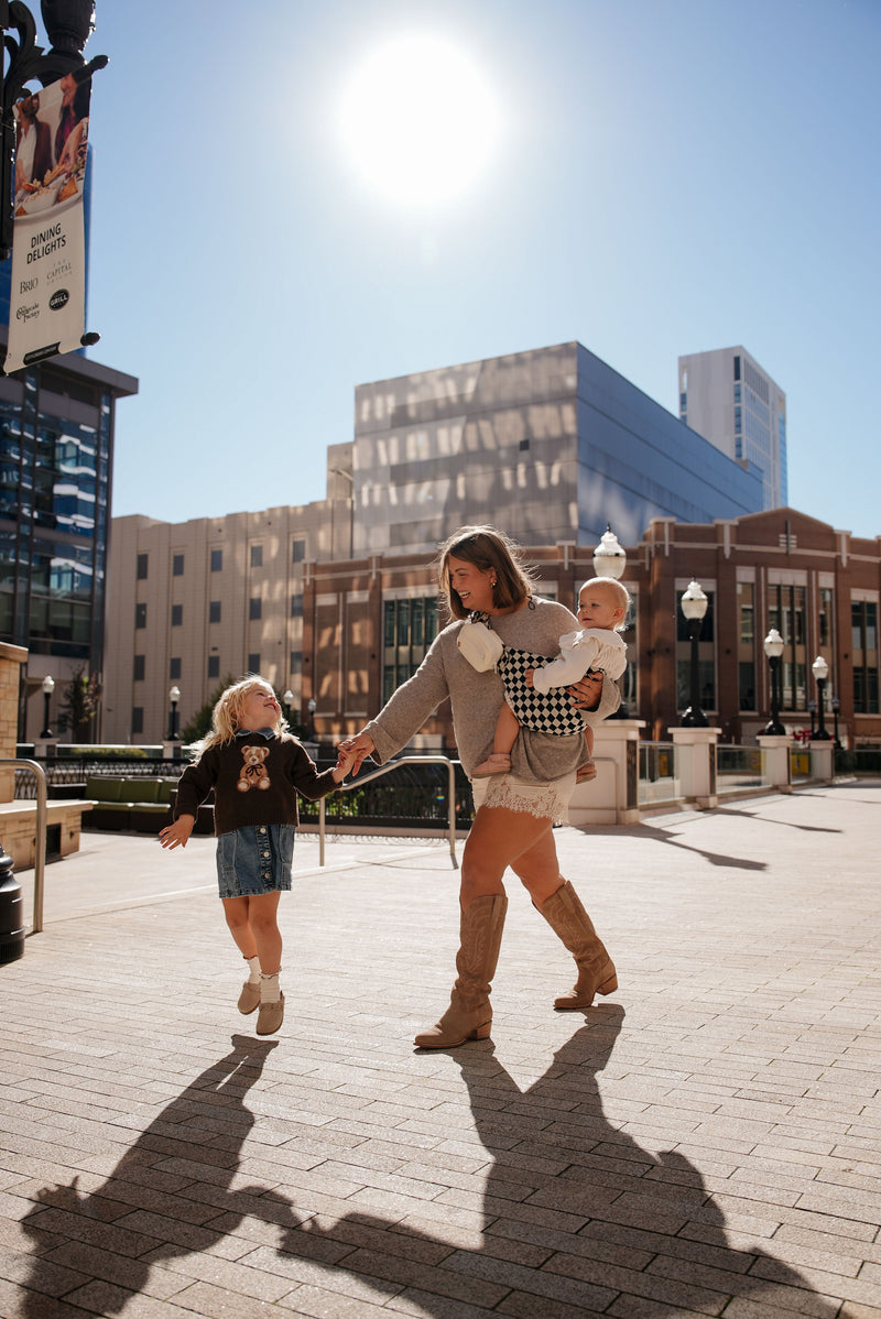 A woman walks outdoors on a sunny day, holding a baby in a black‑and‑white checkered Mabē hip sling while gently swinging the hand of a small child who is mid‑step, hopping with excitement. The woman wears tall tan boots and a light sweater, and the child wears a dark jumper and denim skirt. Long shadows stretch across the brick walkway, and modern city buildings rise behind them, glowing in the bright midday sun.