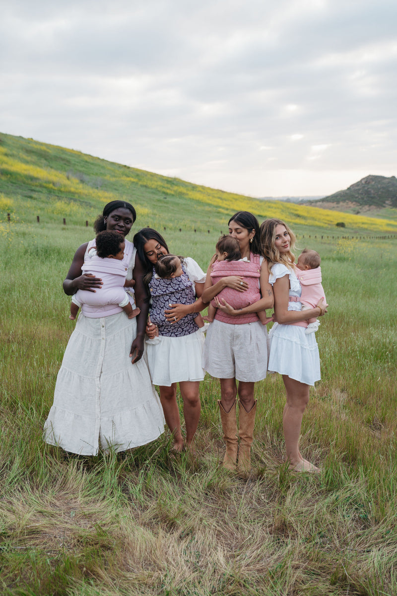 Four women stand together in a grassy, open field surrounded by rolling green hills under a soft, cloudy sky. The first woman has brown skin and curly black hair; the second and third have tan skin and straight black hair. The fourth has light skin with brown wavy hair with some blonde highlights. Each woman is wearing white clothing and holding a young child or baby in a Mabē buckle carrier.