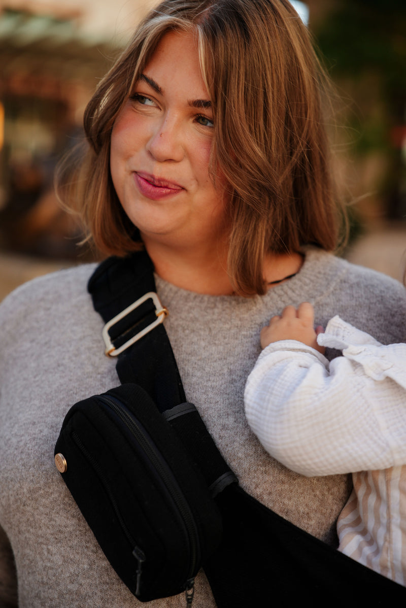 White woman with reddish brown hair carrying a child, who is outside the frame except for their arm, on her hip, with a black Mabē hip sling and accessory bag against a, blurred outdoor background