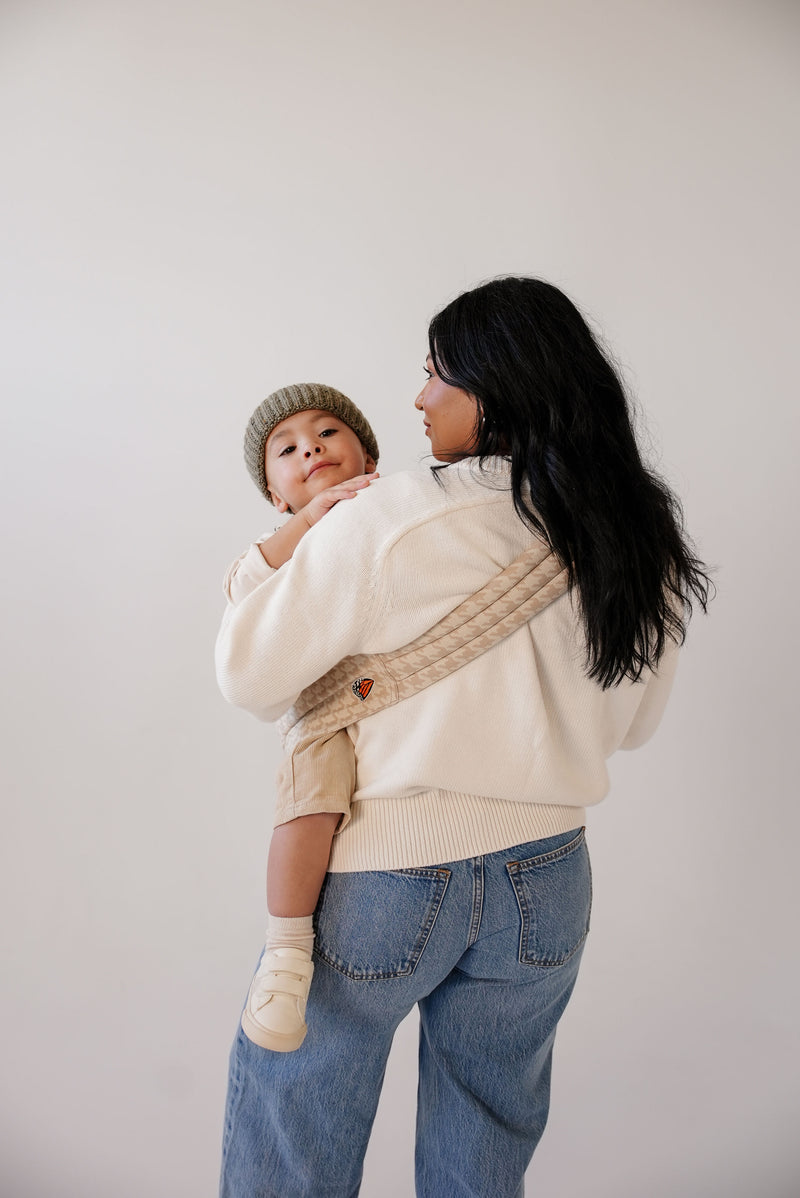 A woman with long wavy black hair stands against a white backdrop, holding a small child on her hip in a beige houndstooth patterned Mabē hip sling. She wears a cream-colored sweater and relaxed blue jeans. The child is dressed in light, neutral tones, including a knit beanie and light socks and shoes. Her long dark hair falls loosely down her back as she faces  away with the toddler peeking over her shoulder, creating a calm, minimal, and cozy scene.