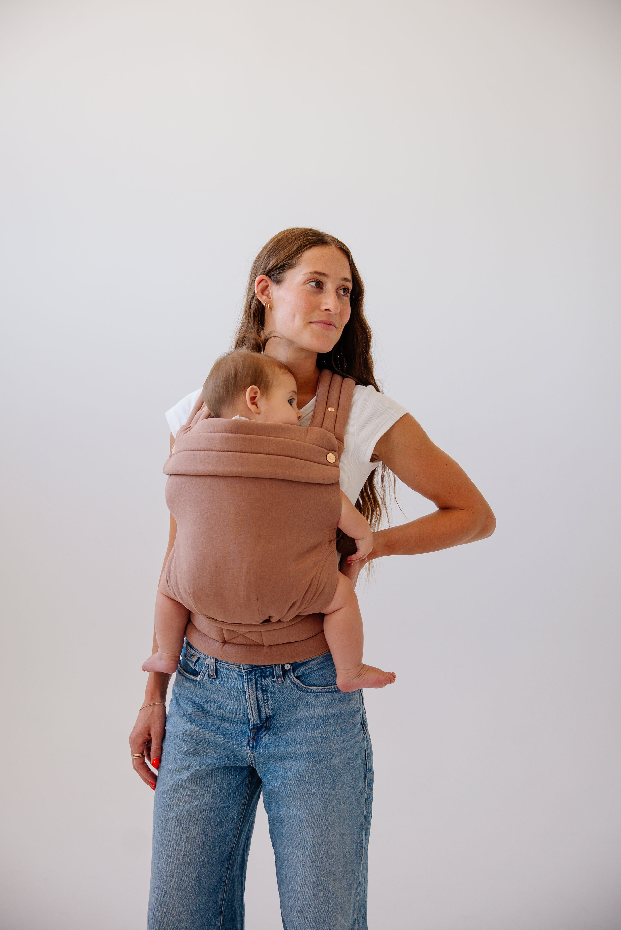 A photo from the knees up of a white woman with long wavy brown hair. She wears mid-wash jeans and a white t-shirt and a baby with light brown hair in a solid mocha-colored Mabē Monarch buckle carrier. She looks off to the right of the frame with her hand on her hip.