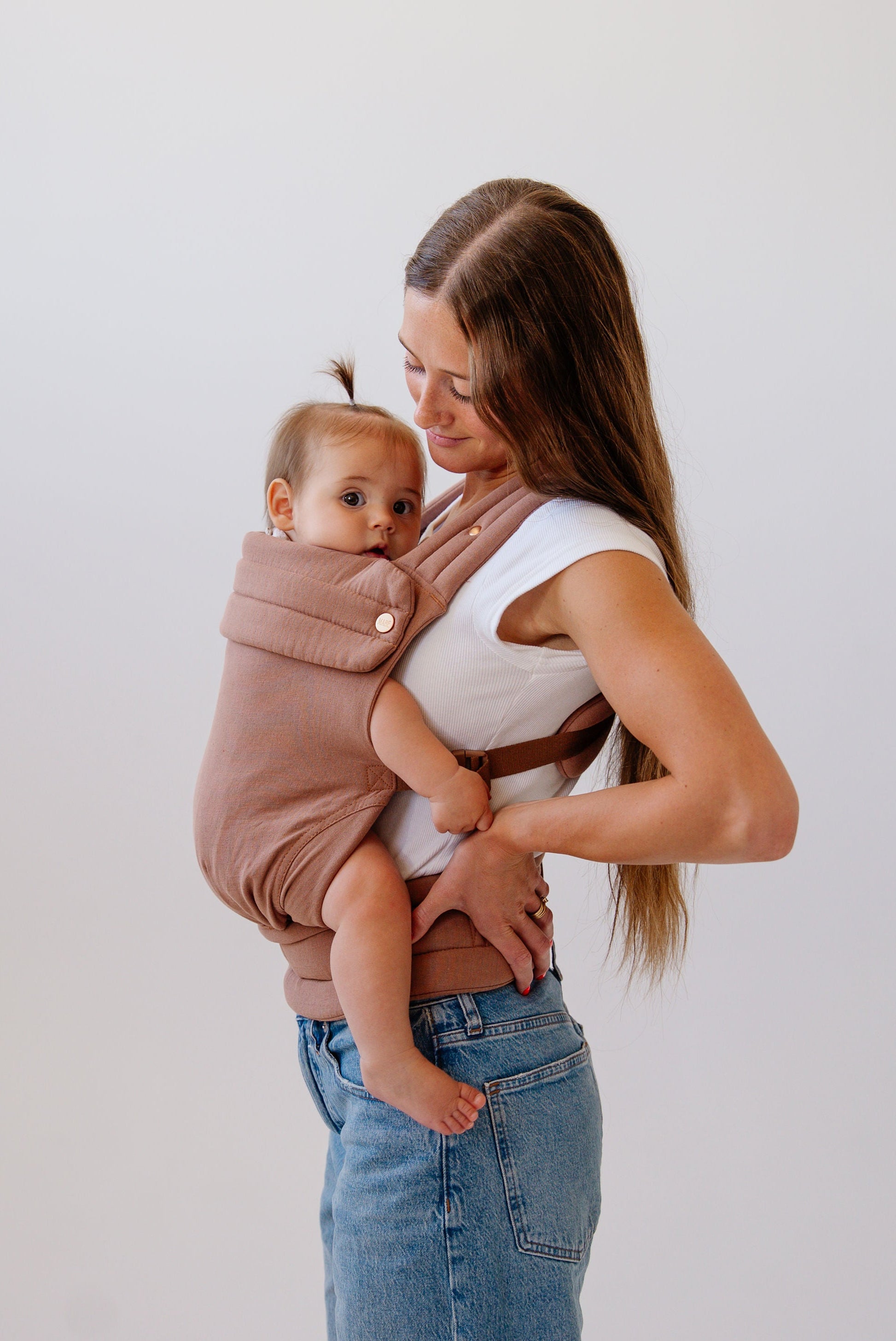A photo from the knees up of a white woman with long wavy brown hair standing sideways facing the left of the frame. She wears mid-wash jeans and a white t-shirt and a baby with light brown hair and a tiny ponytail on the top of their head in a solid mocha-colored Mabē Monarch buckle carrier. She looks down at the baby in the carrier and has her hand on her hip.