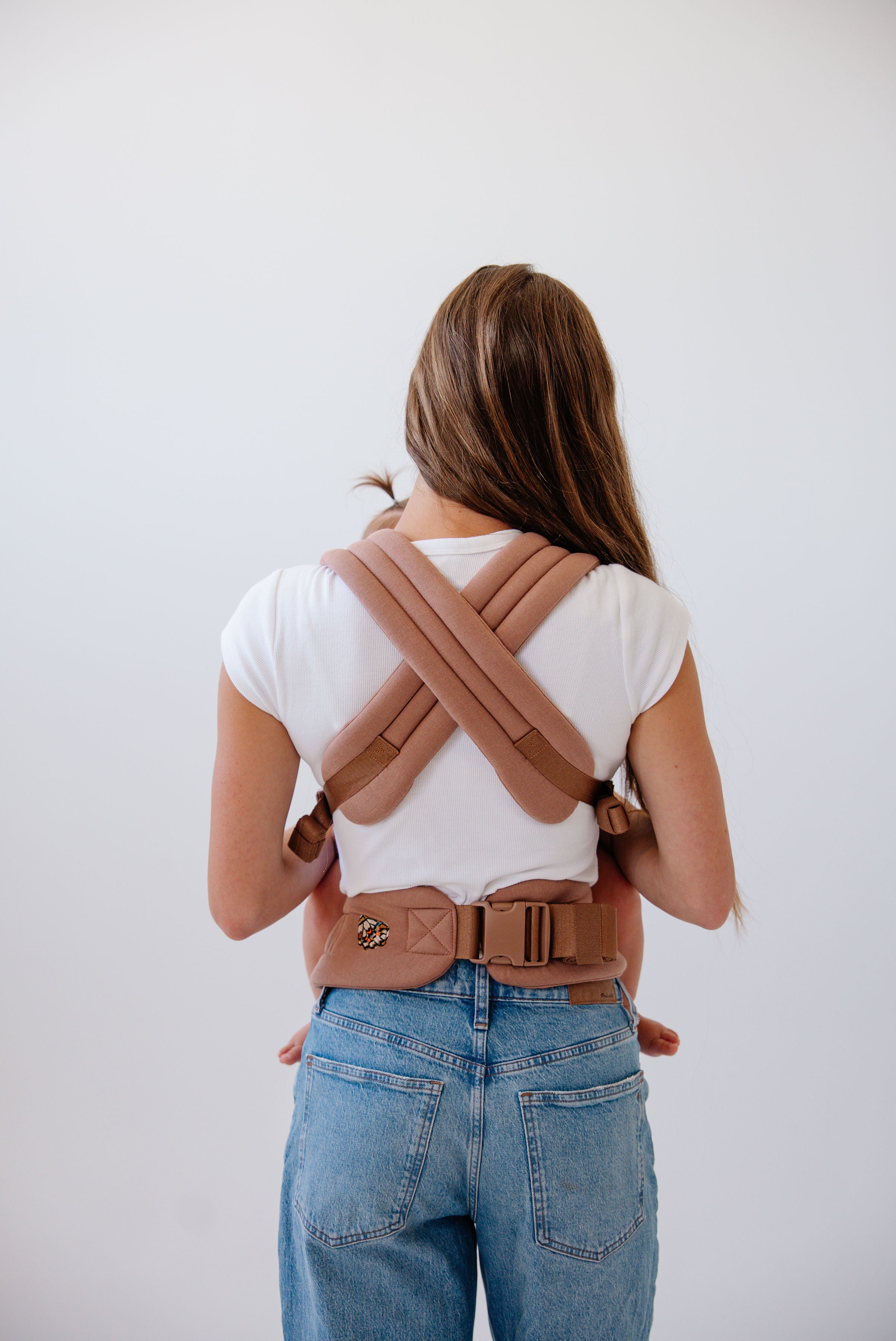 Photo from the knees up of a woman with long brown hair facing away from the camera. She wears mid-wash jeans and a white t-shirt and has the solid mocha Mabē Monarch buckle carrier on with a mostly unseen baby in it. The straps cross in an "X" shape across her back and the waistband clips at the small of her back. A tiny baby ponytail is visible above her left shoulder and baby toes are visible next to her hips. 