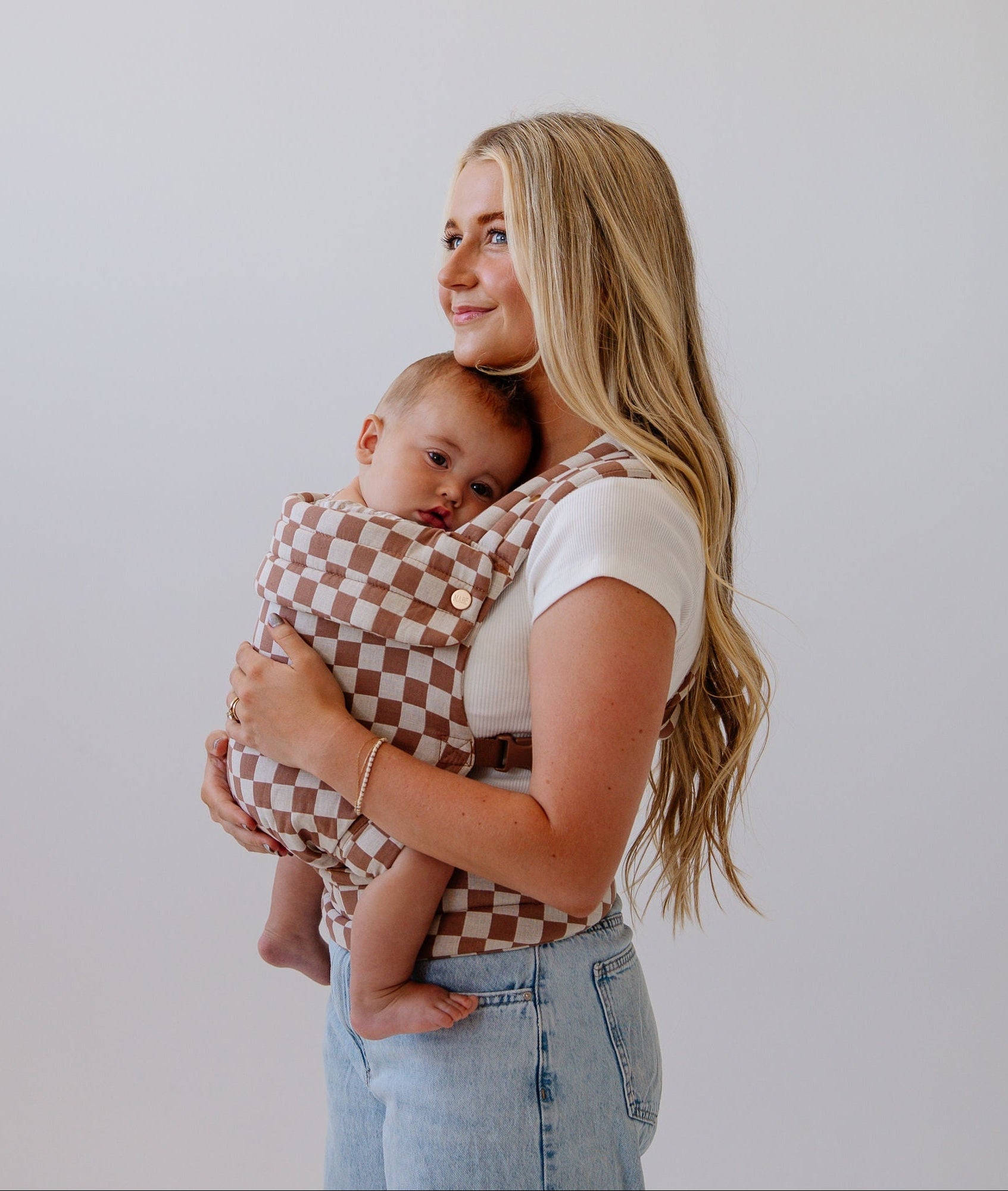 A tanned white woman with long blonde highlighted hair in a white t-shirt and light wash jeans stands sideways looking to the left of the frame, against a white background, wearing a sleepy baby in a checkered carrier in cream and pinky brown mocha.