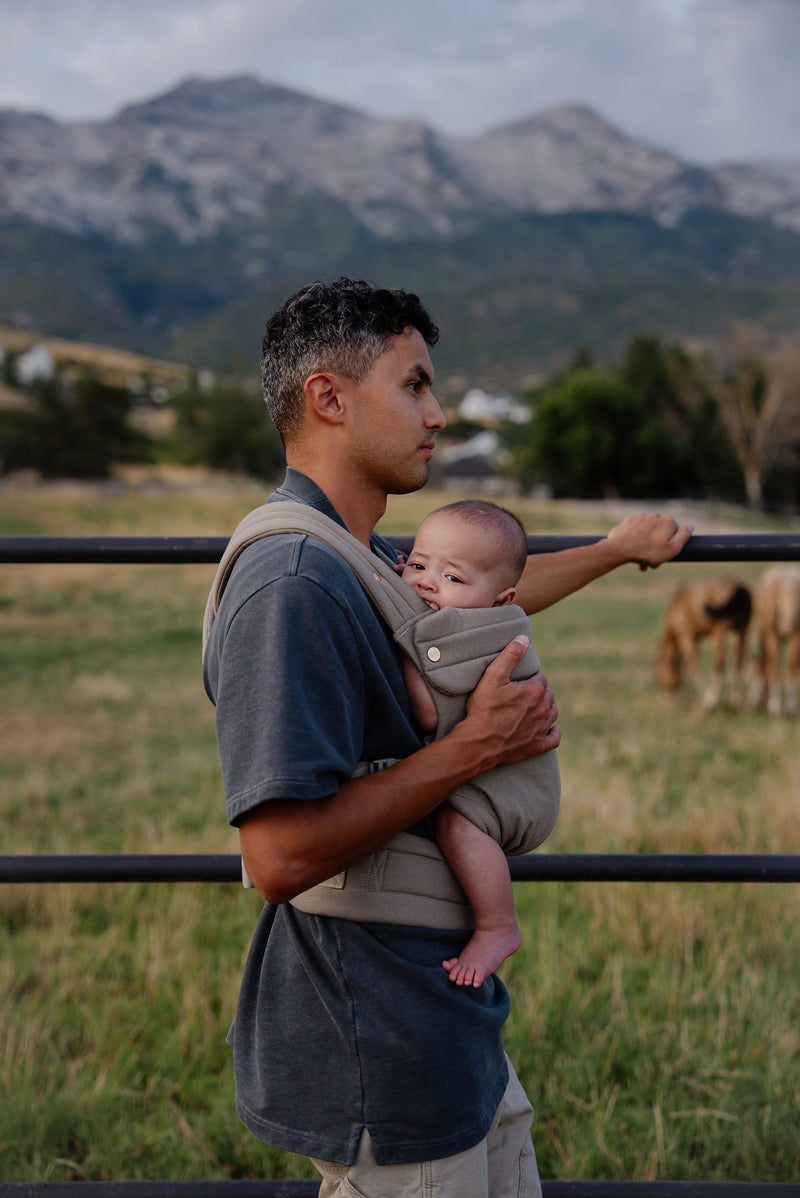 A dark-haired, tan-skinned man stands in a grassy field near a metal fence, holding a baby snugly against his chest in a soft sage-colored Mabē buckle carrier. He rests one arm on the fence while the other supports the child, whose bare feet peek out from the carrier. Behind them, the landscape opens into rolling fields with a few grazing animals and tall trees, and a mountain range rises in the distance under a cloudy sky. The photo has a modern rustic vibe.