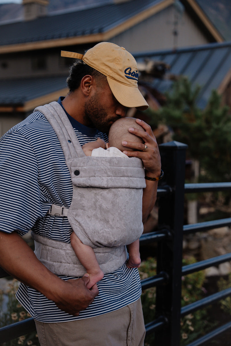 A brown-skinned man with black hair and beard stands outdoors near a black metal railing, holding a baby close to his chest in a soft, light‑gray Mabē carrier. He wears a striped shirt, tan pants, and a beige baseball cap. The baby’s small legs dangle gently from the carrier as the man cradles the child with one hand and presses a tender kiss to the top of the baby’s head. Behind them, a rustic building and trees create a warm, early‑evening atmosphere.