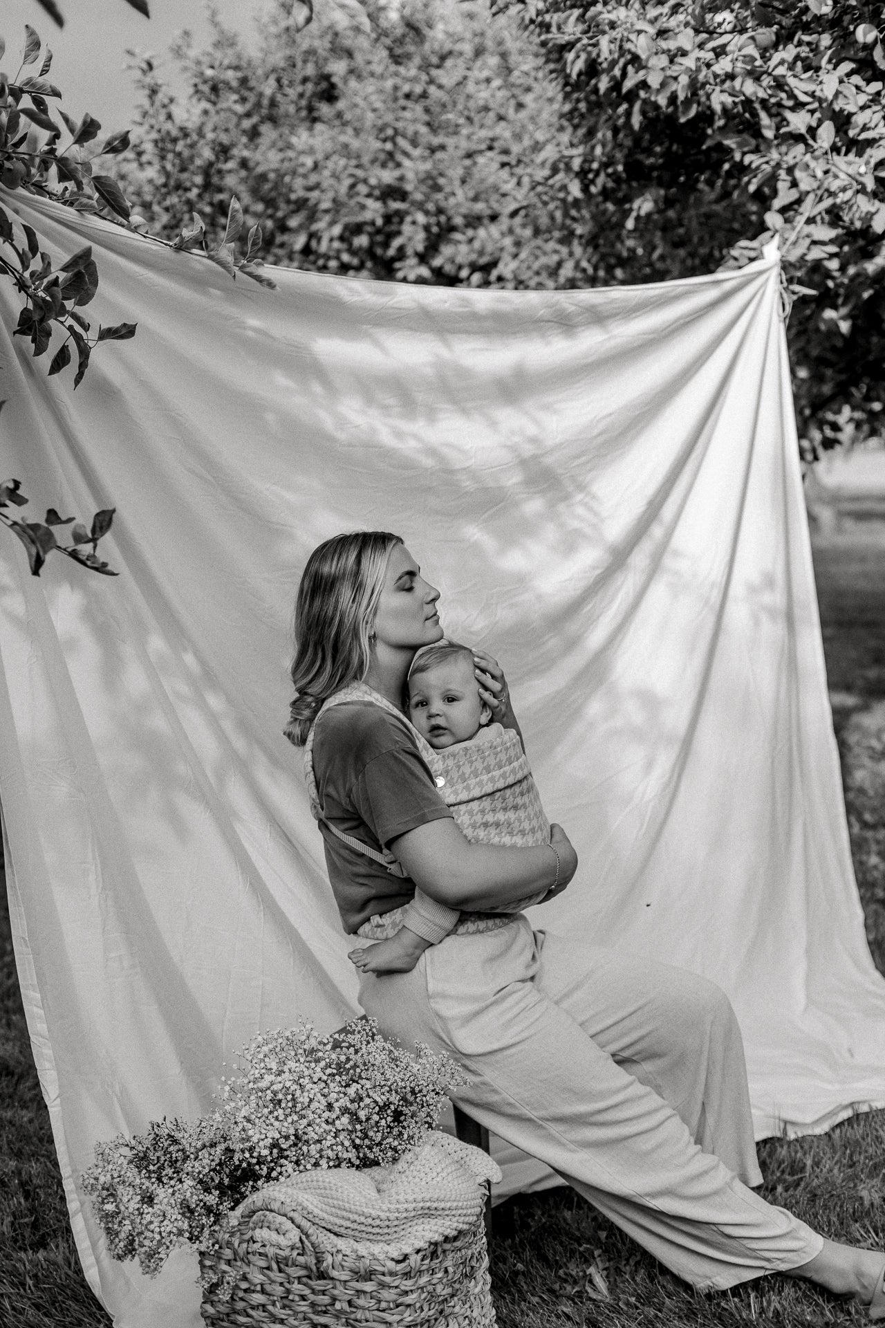 A black-and-white photo of a woman with long hair seated and facing the right of the frame, holding an alert baby in the Mabē Monarch Buckle Carrier with a houndstooth pattern. She is in front of a white backdrop strung between the boughs of two tress, and a basket of bunches of tiny flowers and a knit or waffle blanket is just adjacent to her.