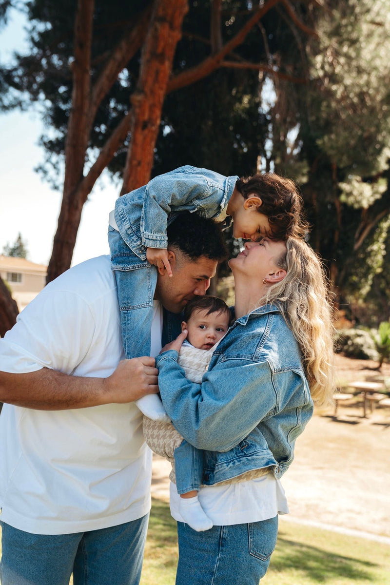 Family of four outdoors in white and denim attire with a dark-haired child in denim pants and jacket, playfully climbing on the tan-skinned, dark-haired man's shoulders. The child is leaning over to kiss his light-skinned, blonde-haired mama, who is holding another younger child in a beige houndstooth patterned Mabē baby carrier.