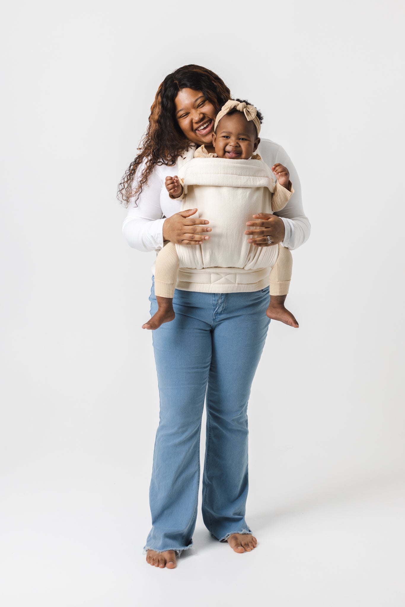 A full-length photo of a brown-skinned woman with long dark curly hair with caramel highlights carrying a young toddler in the Mabē Monarch Buckle Carrier in Natural Ivory, which is solid cream colored. The baby is positioned world-facing, and is grinning. She wears cream-colored leggings and bow. The woman wears a white long-sleeved shirt and light-wash denim jeans.