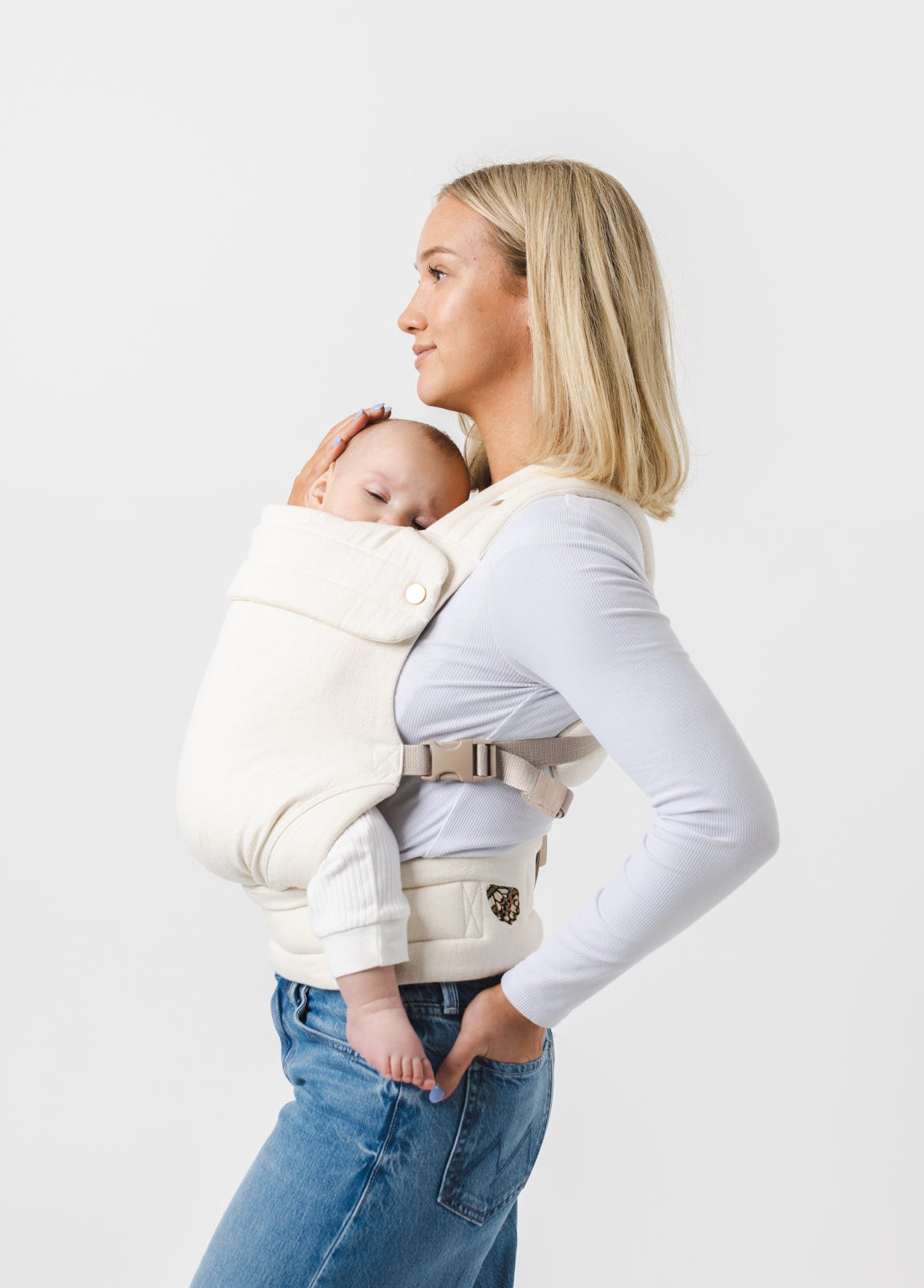 Photo from above the knees up of a white woman with shoulder-length blonde hair standing sideways looking to the left of the frame. She is wearing a long-sleeved white shirt and mid-wash jeans and carries a sleeping baby in an ivory-colored Mabē Monarch buckle carrier.