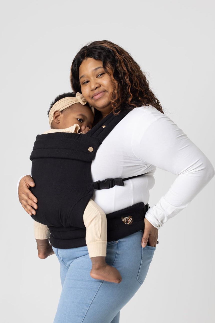 Photo from the knees up of a tan-skinned woman with highlighted black hair standing sideways looking at the camera. She wears a long-sleeved white shirt and light-wash jeans and carries a black-haired baby with a beige headband in a solid black Mabē Monarch buckle carrier.