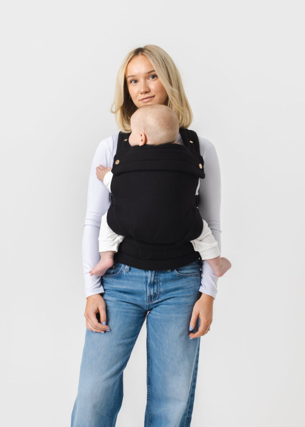 A woman with long bobbed blonde hair and tanned skin faces the viewer. She is shown from the knees up. She is wearing wide leg medium wash jeans and a long sleeved white fitted shirt and a Mabē Monarch Buckle Carrier in Ebony, which is solid black. She looks at the viewer. The baby has long cream colored pants and shirt on. The woman has light blue nails.