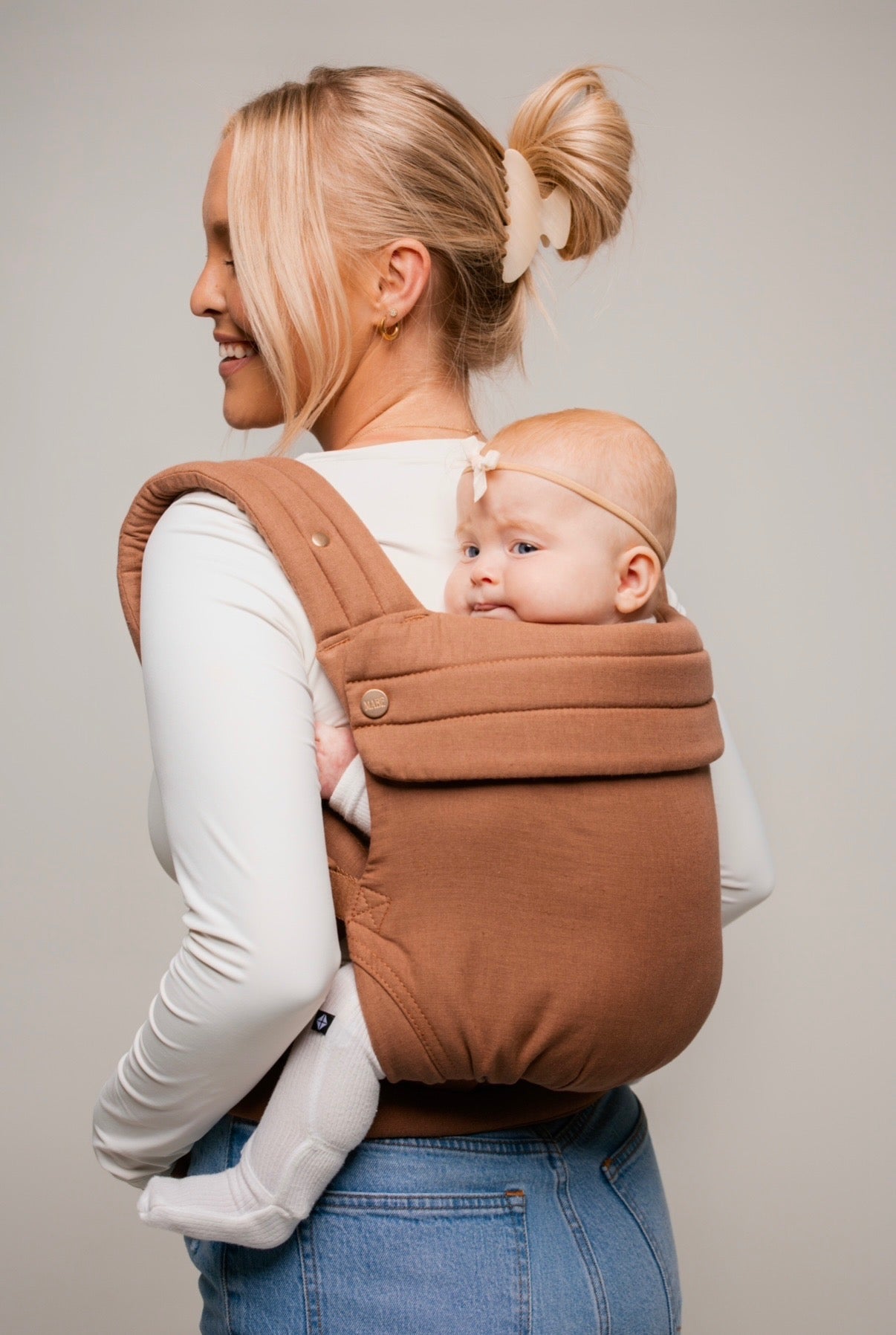 Photo from the hips up of a white woman in a long-sleeved white shirt and mid-wash jeans standing sideways, almost with her back to the viewer, wearing a bald baby with a thin headband with a tiny bow in a pinky-brown solid Mabē Monarch mocha buckle carrier. Her hair is clipped up in a loop with a beige claw clip and has long layers framing the visible side of her face.