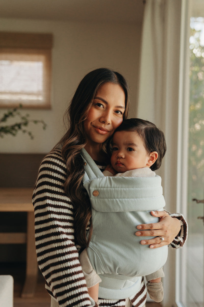 A dark-haired woman is standing indoors holding a young child close to her chest in a soft, light‑blue Mabē Buckle baby carrier. She has long, wavy brown hair and is wearing a cozy, striped sweater with dark and light brown tones. The child is nestled comfortably against her, with tiny arms and legs partially visible, dressed in neutral‑colored clothing and socks. The background shows a warm, homey room with soft natural light, a wooden table, and light-colored curtains, creating a calm atmosphere.