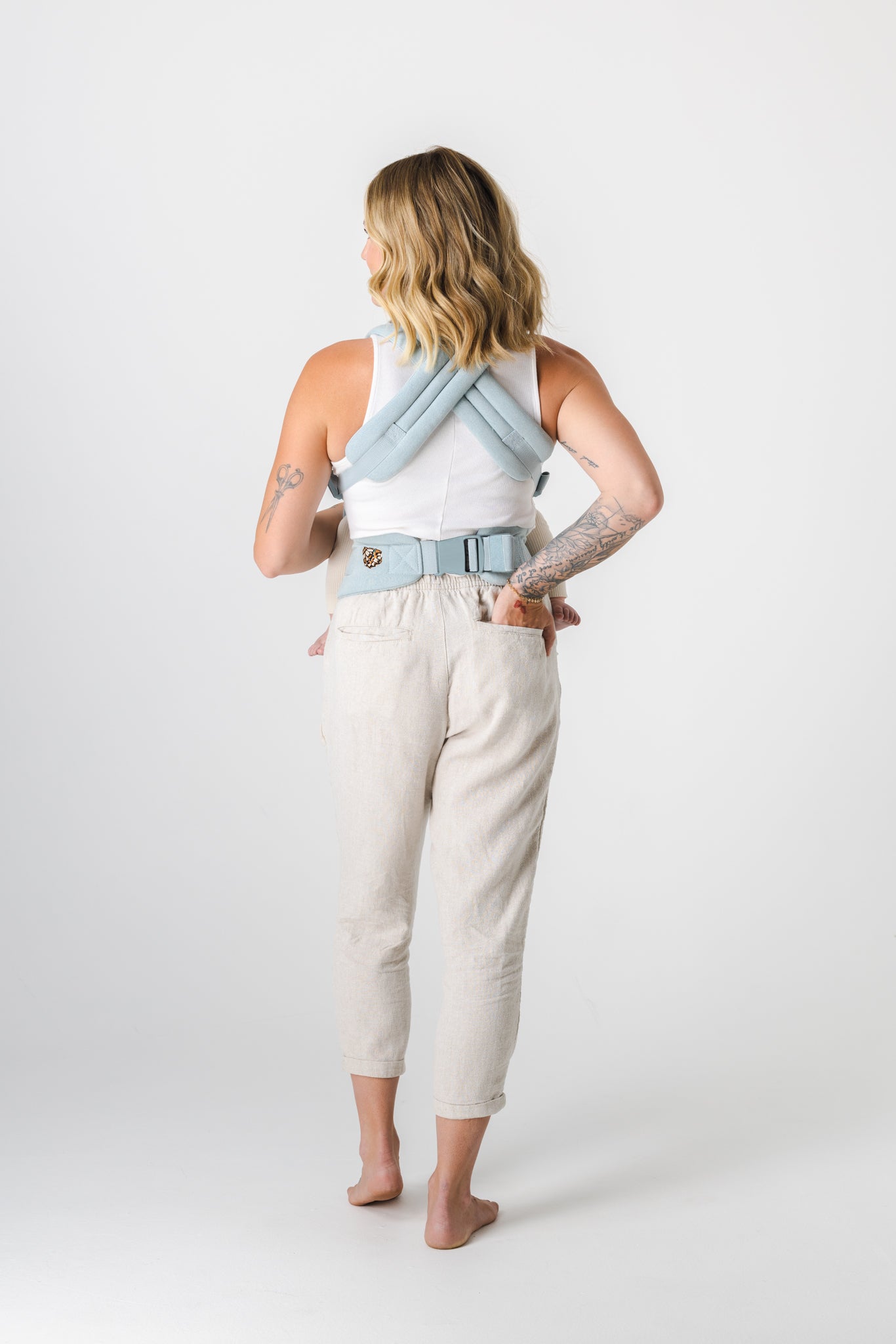 Full-length photo from the back of a person with an unseen face and curled light brown hair with blonde highlights. They are wearing a white racerback tank top and cream colored culottes, and the Mabē Monarch Buckle Carrier in Dream Wash Denim, which is solid light blue in a denim effect cotton fabric. Their right hand is in their back pocket and you can jut barely tell they are looking slightly to their left. They are barefoot and the background is solid white.