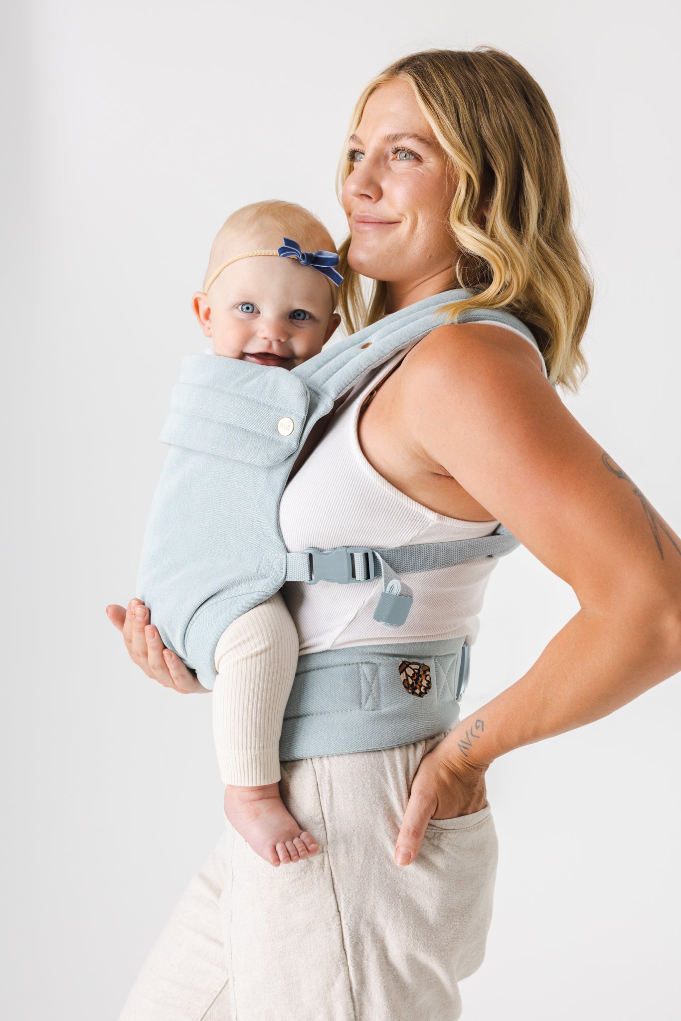 A woman with curled shoulder-length light brown hair with blonde highlights faces the left of the frame. She is shown from the mid-thigh up and is wearing a white tank top and cream-colored bottoms. She has a closed-lipped smile and wears the Mabē Monarch Buckle Carrier in Dream Wash Denim, which is solid light blue in a cotton denim effect fabric. There is a young toddler girl in the carrier with light skin and hair wearing cream-colored pants. She looks at the viewer with a smile. The background is white.