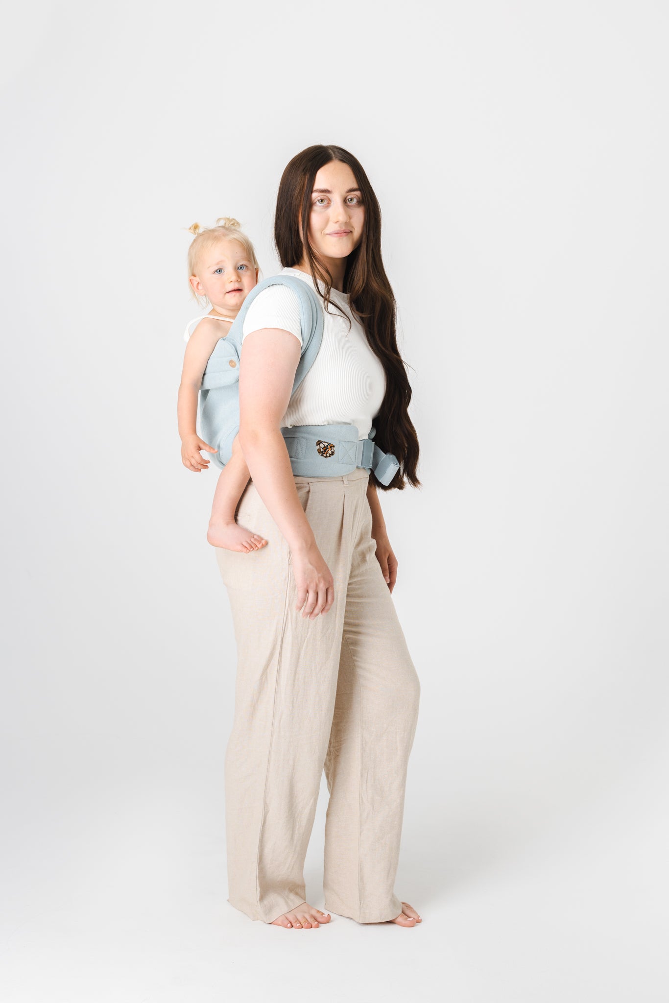 A full-length photo of a light-skinned woman with waist-length brown hair wearing a white t-shirt and long wide-leg cream colored trousers. She is wearing on her back the Mabē Monarch Buckle Carrier in Dream Wash Denim  with a light-skinned, very blonde toddler girl in the carrier. They both have slight smiles. The background is solid white.