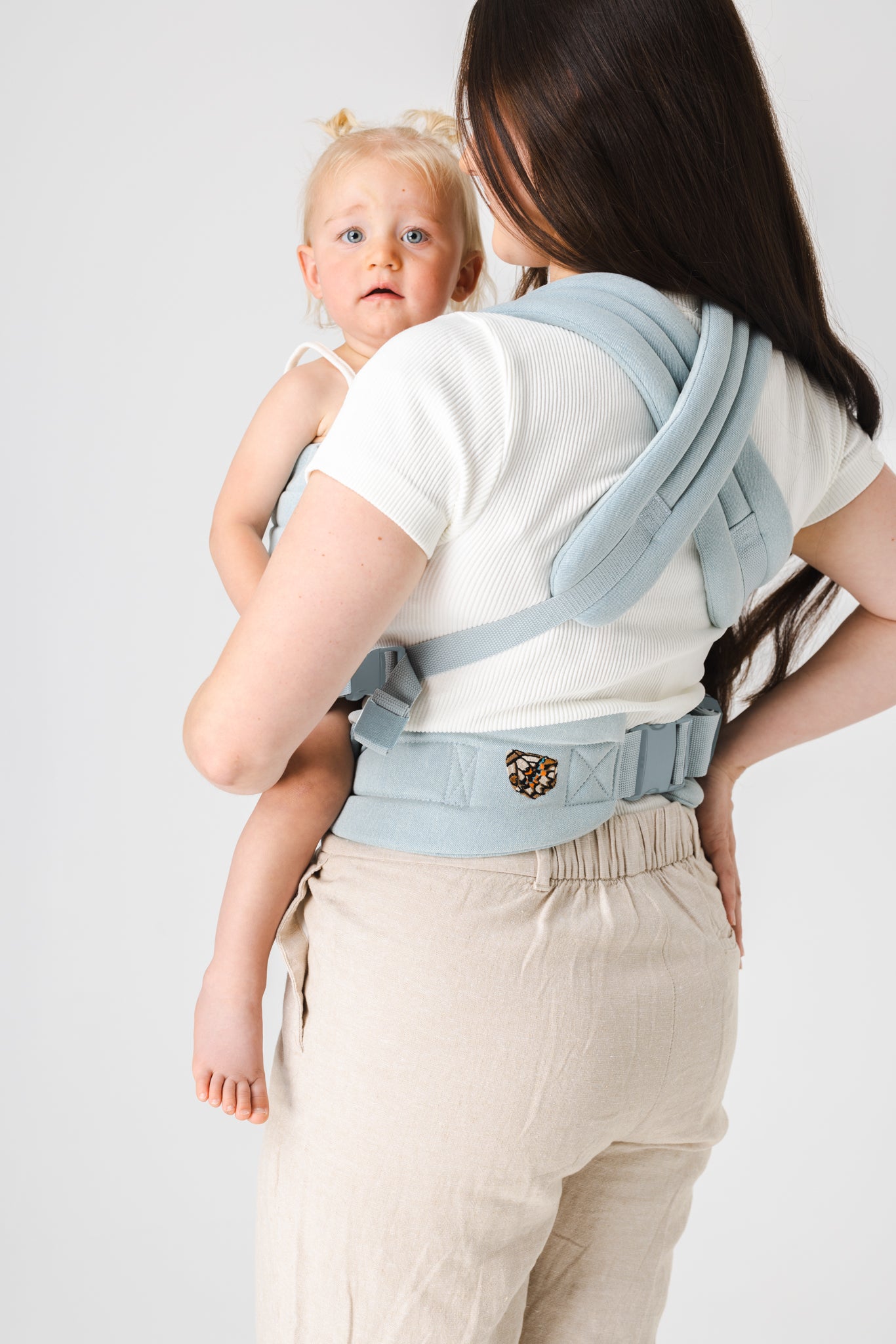 Back view of a person with waist-length brown hair wearing the Mabē Monarch Buckle Carrier in Dream Wash Denim, which is solid light blue with a light wash denim jeans effect. They are wearing the carrier on their front, and the blonde toddler girl in the carrier looks over the shoulder of the wearer with a cute, inquisitive expression on her face. The background is solid white.