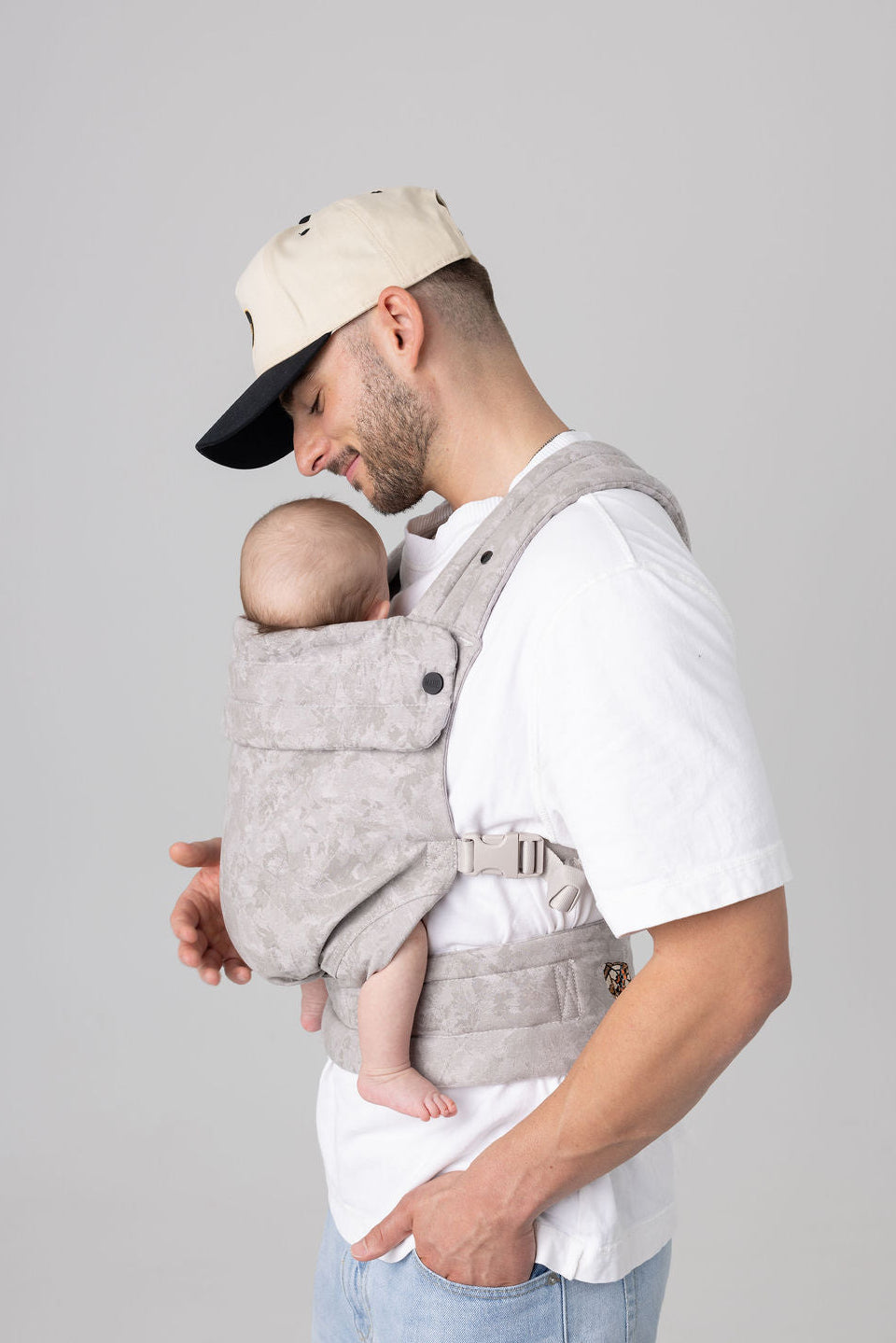 Side view of a white man with dark facial hair. He wears a tan ball cap with a black brim and carries a light-skinned baby in a light-colored, warm-gray camouflage-patterned Mabē Monarch Buckle carrier. The colorway is Ashland Camo. The man wears a white t-shirt and light-wash denim. He is shown from the mid-thigh up.