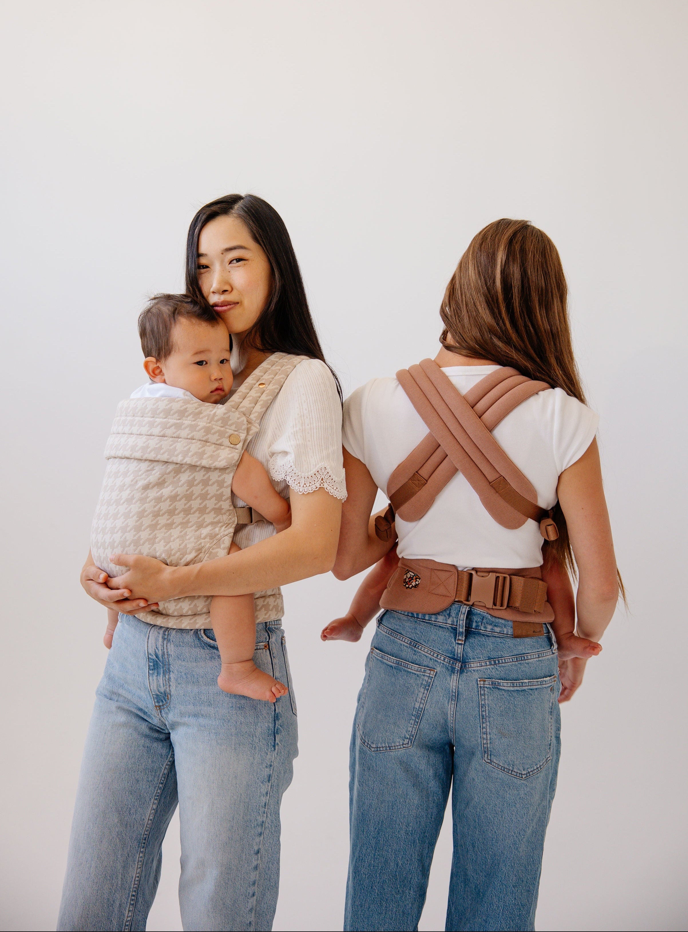 Two women stand in a bright, minimal studio, each wearing a Mabē Buckle carrier. One faces forward holding a child in a houndstooth-patterned beige carrier, while the other stands turned away, showing the crossed straps and waistband of a rosewood‑colored carrier. Both wear casual white tops and blue jeans, highlighting the carriers’ fit and design.