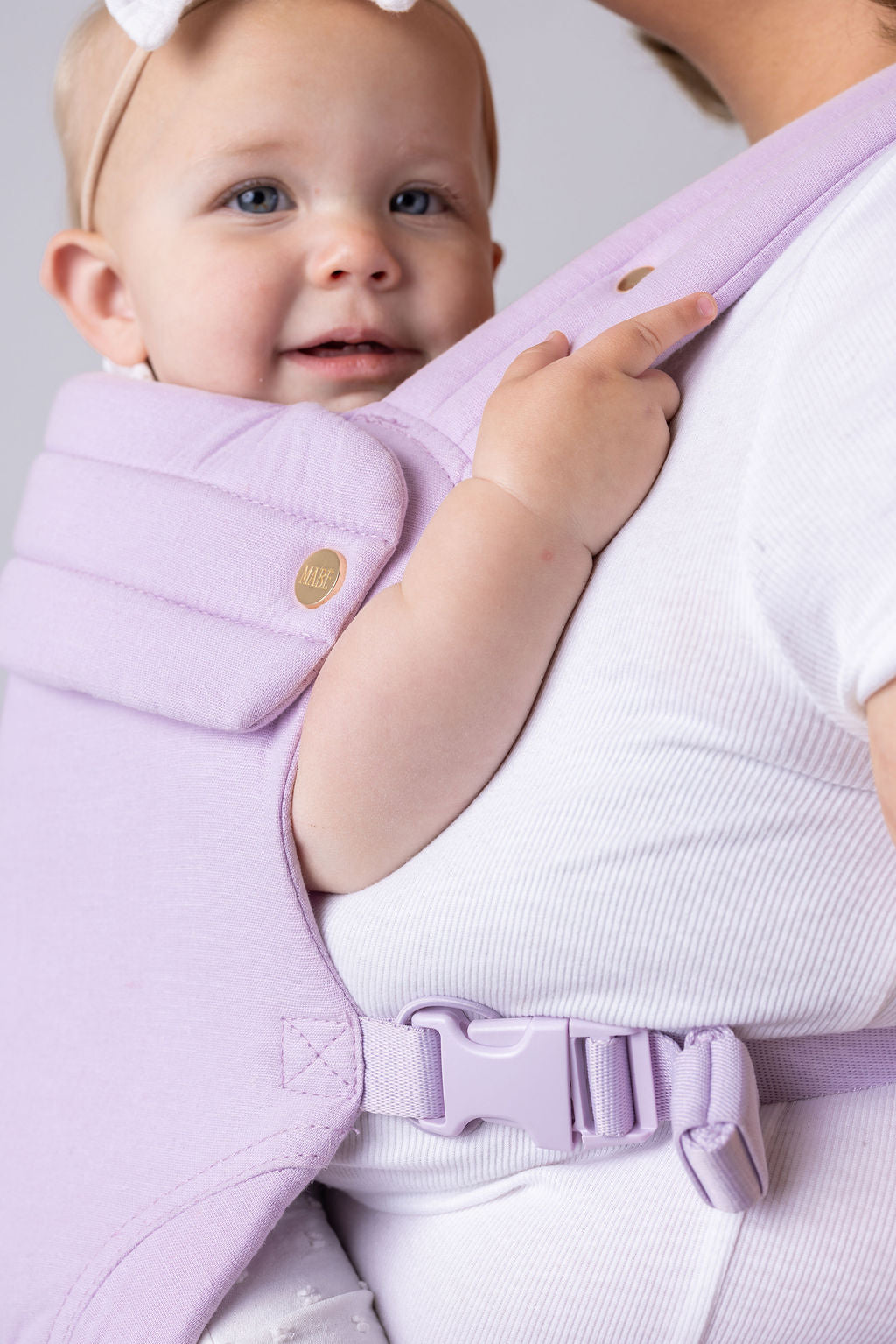 Close-up photo of an unseen woman featured in other photos. She is wearing a light-skinned baby with a white bow in the Mabē Monarch Buckle Carrier in Lilac Skies which is light purple. She is shown from the waist up and chin down.