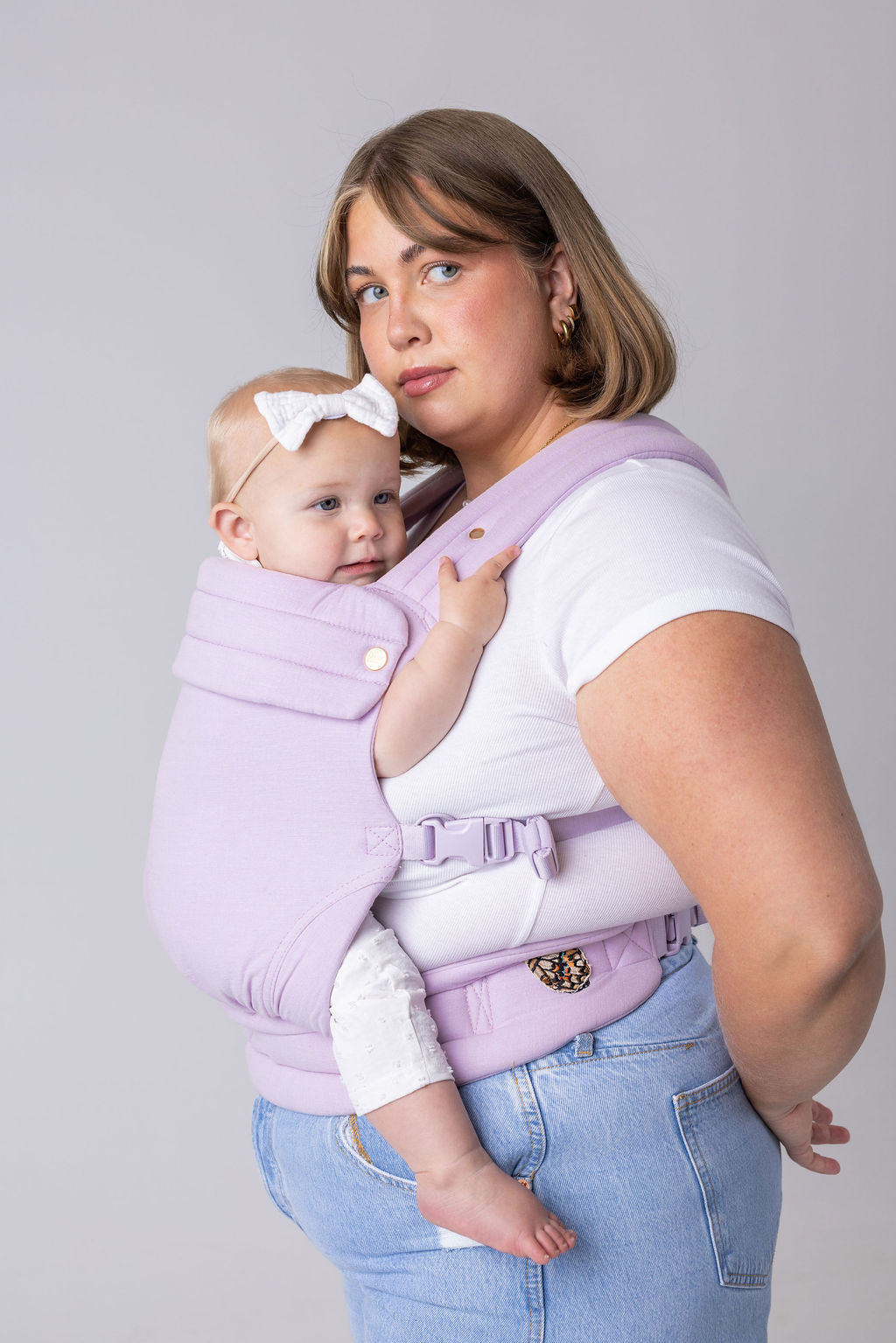 A light-skinned woman with bobbed dark blonde hair faces the left of the frame. She wears a white t-shirt and light wash jeans. She is wearing a light-skinned baby with a white bow in the Mabē Monarch Buckle Carrier in Lilac Skies which is light purple. She is shown from the bottom up.
