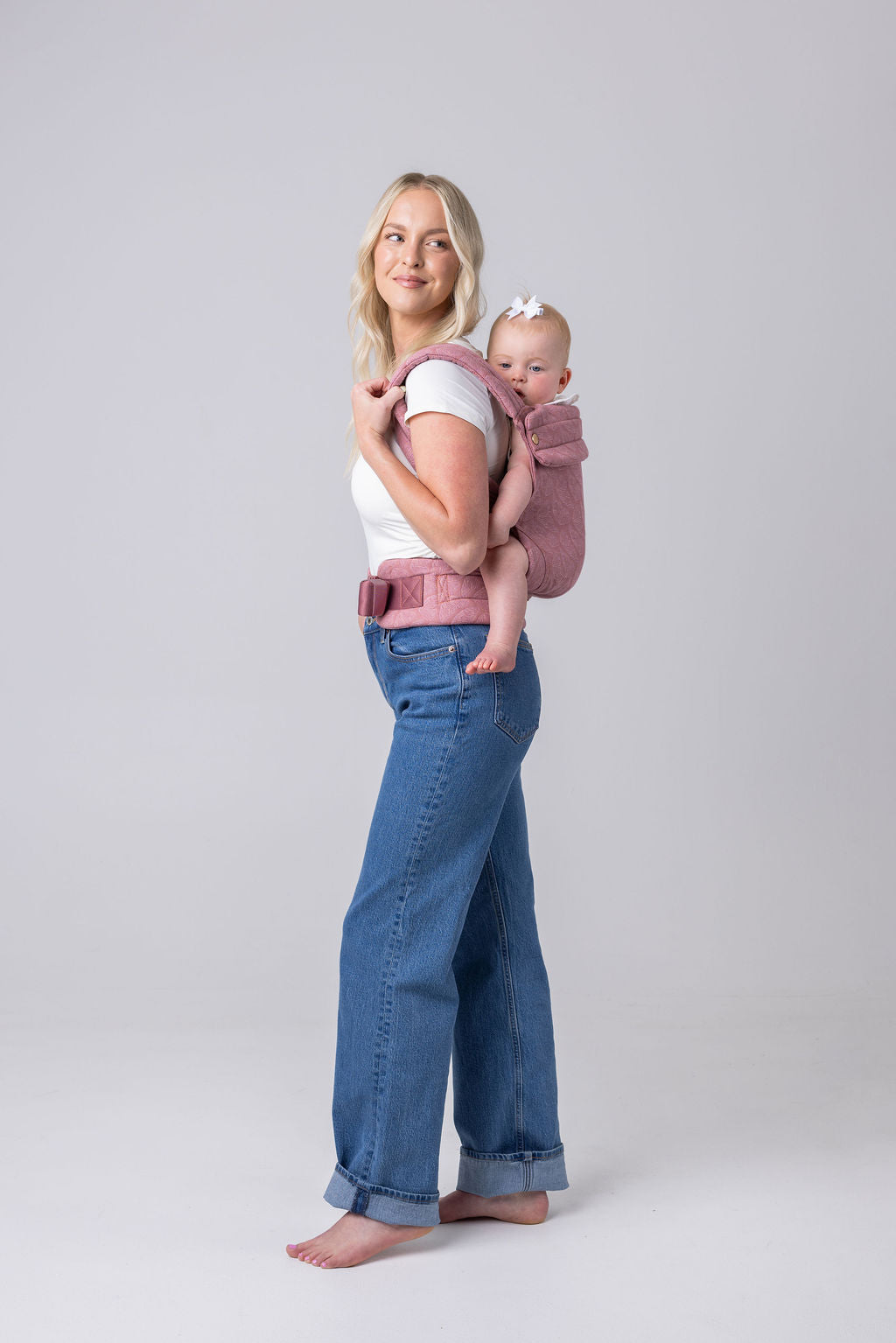 A light-skinned woman with long blonde hair, a white t-shirt, and medium-wash jeans carries a light-skinned, light-haired baby in the Mabē Monarch Buckle carrier in Mauve Wings, which is blush pink with lighter butterfly wings patterned on it. She faces the left of the frame, is looking to the right of the frame, and is shown full height. The background is white or light gray. She is barefoot.