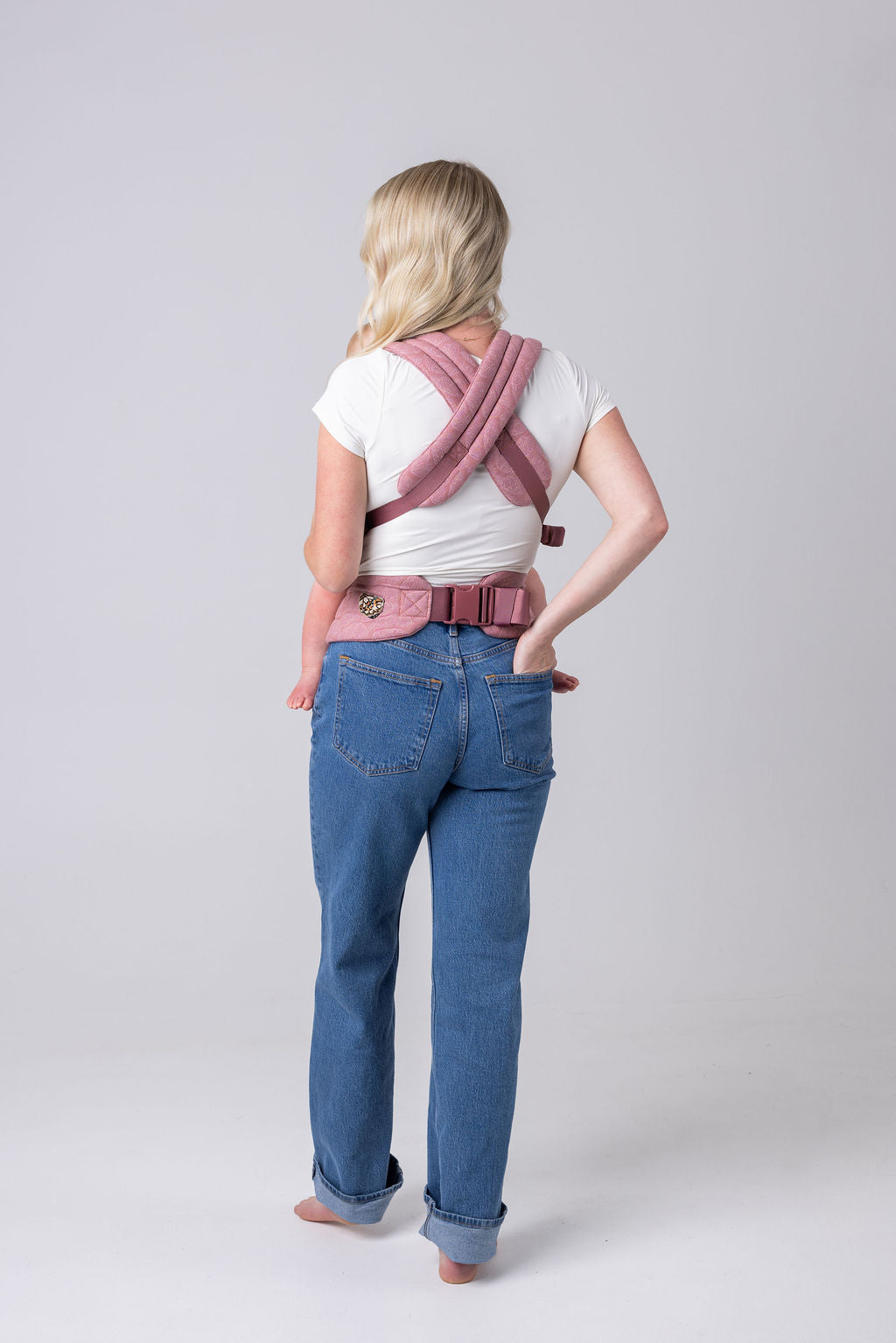 A person with an unseen face and long blonde hair faces away from the viewer. They have the crossed straps of the Mabē Monarch Buckle carrier in Mauve Wings across their back and the waistband buckled around their waist. They wear a white t-shirt and medium wash jeans. They are barefoot.