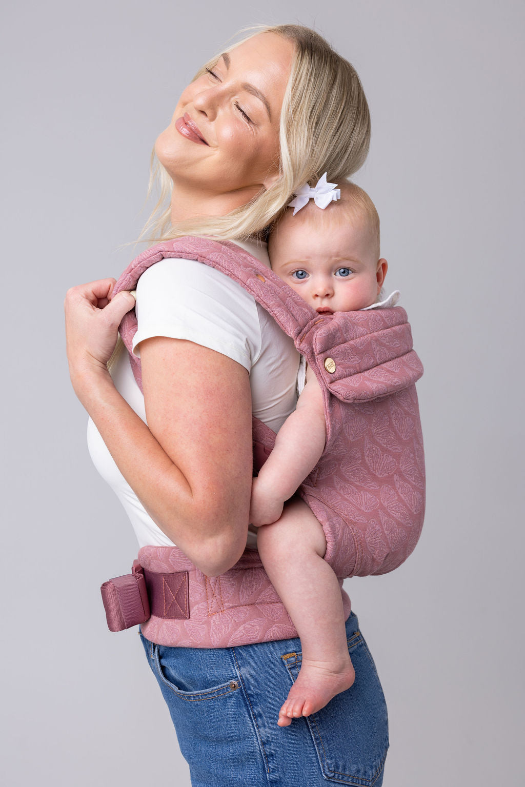 A photo of a light-skinned woman with long blonde hair, a white t-shirt, and medium-wash jeans. She carries a light-skinned, light-haired baby on her back in the Mabē Monarch Buckle carrier in Mauve Wings, which is blush pink with lighter butterfly wings patterned on it. She faces the left of the frame and is tilting her head back to touch the baby's. The background is white or light gray.