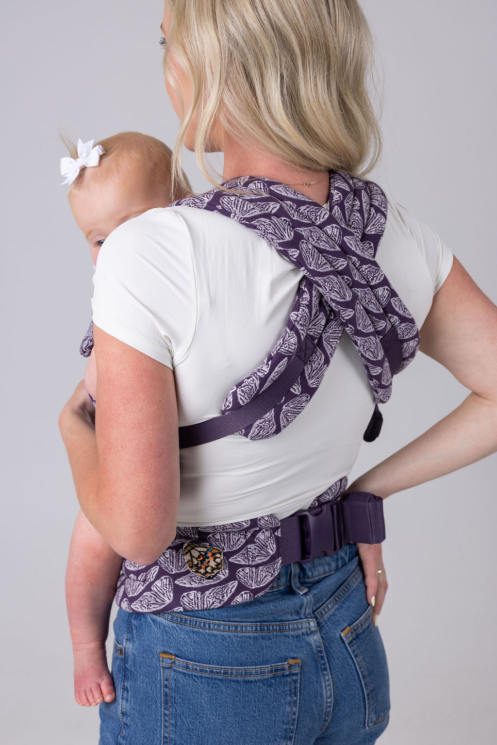 A light-skinned woman with long blonde hair, a white t-shirt, and medium-wash jeans carries a light-skinned, light-haired baby in the Mabē Monarch Buckle carrier in Violet Wings, which is dark purple with lighter butterfly wings patterned on it. She faces away from the frame and is shown from the bottom up with the carrier straps crossed across her back and the waistband buckled on. The background is white or light gray.