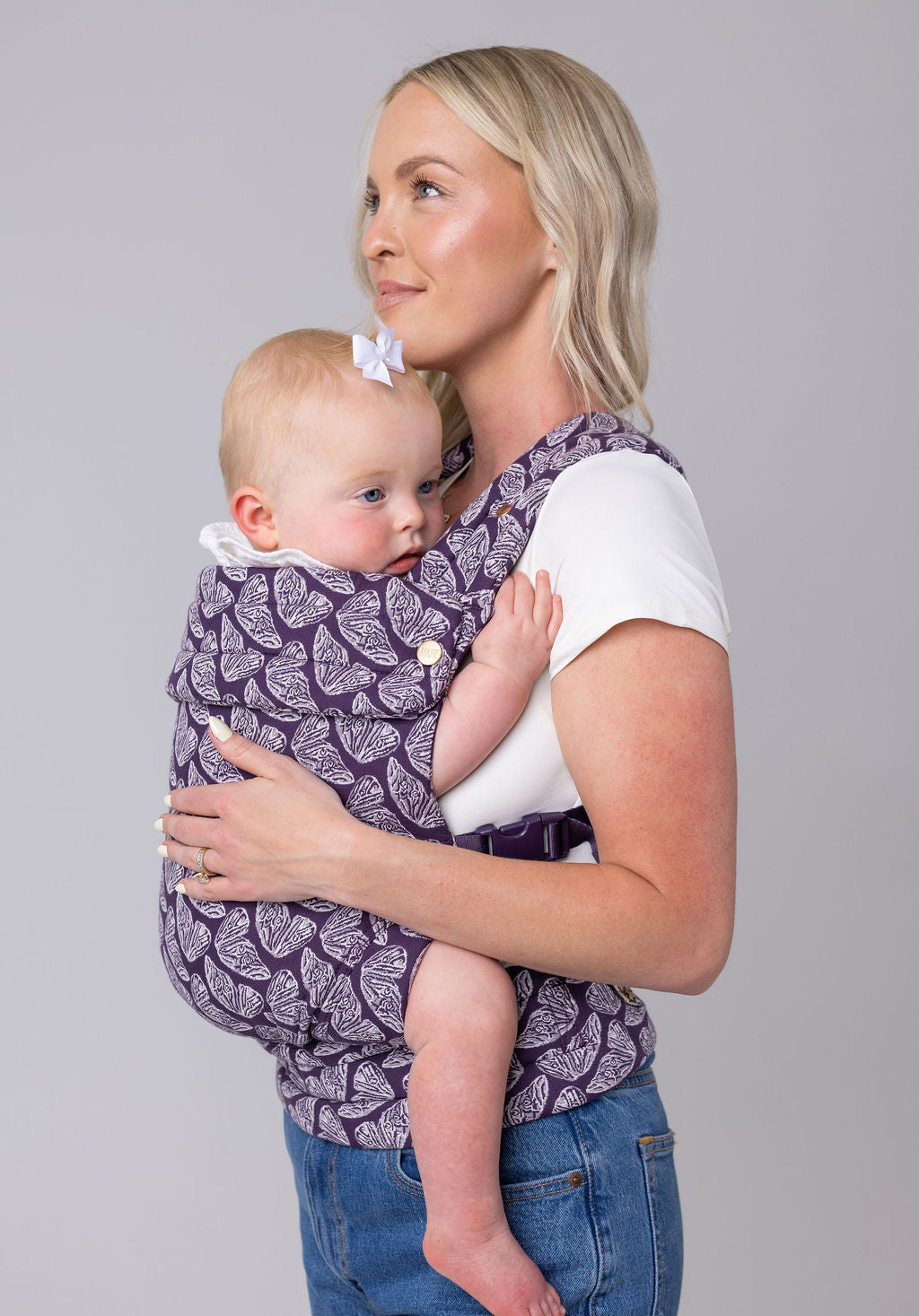 A light-skinned woman with long blonde hair, a white t-shirt, and medium-wash jeans carries a light-skinned, light-haired baby in the Mabē Monarch Buckle carrier in Violet Wings, which is dark purple with lighter butterfly wings patterned on it. She faces the left of the frame and is shown from the hips up. The background is white or light gray.