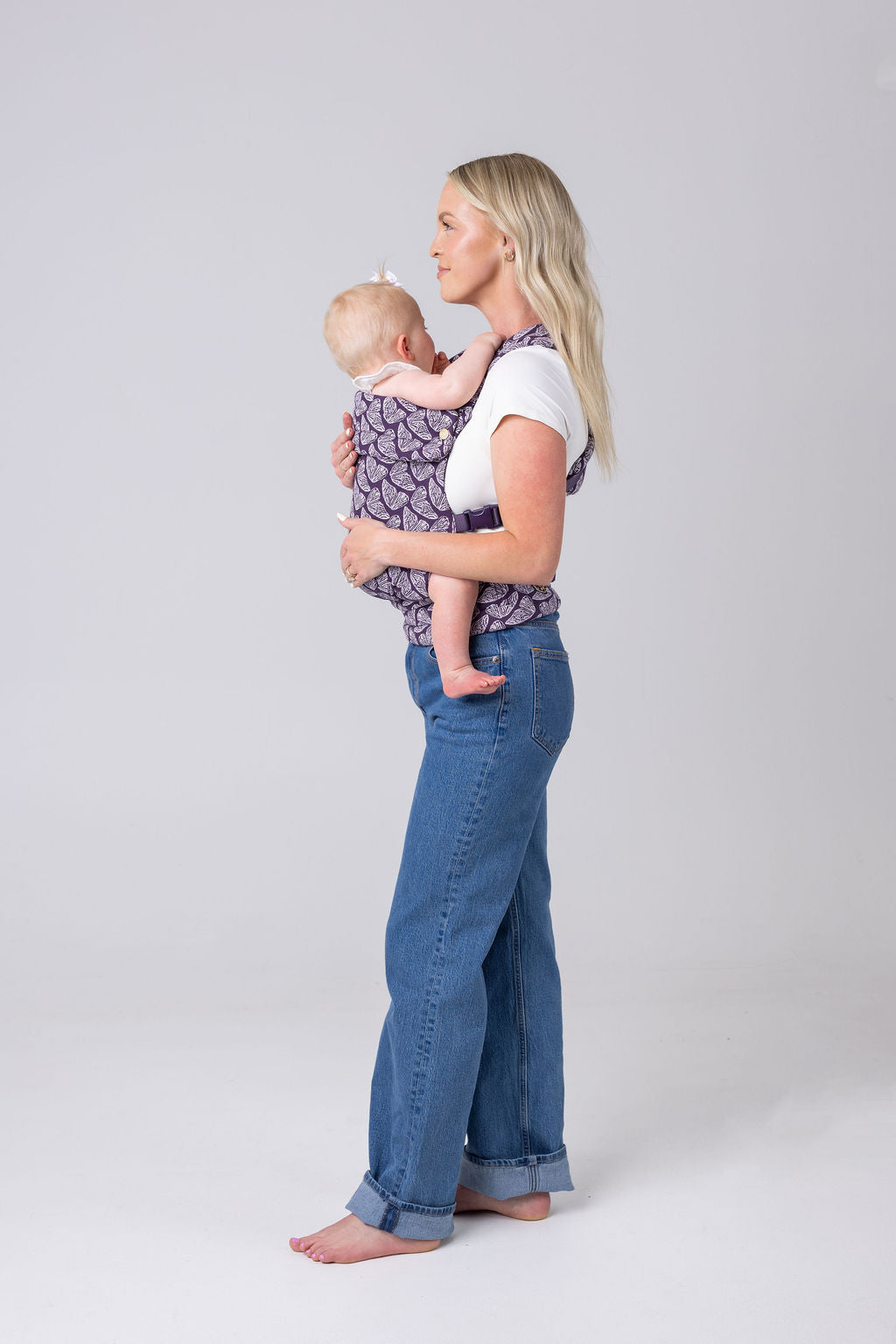 A light-skinned woman with long blonde hair, a white t-shirt, and medium-wash jeans carries a light-skinned, light-haired baby in the Mabē Monarch Buckle carrier in Violet Wings, which is dark purple with lighter butterfly wings patterned on it. She faces the left of the frame and is shown full height. The background is white or light gray. She is barefoot.