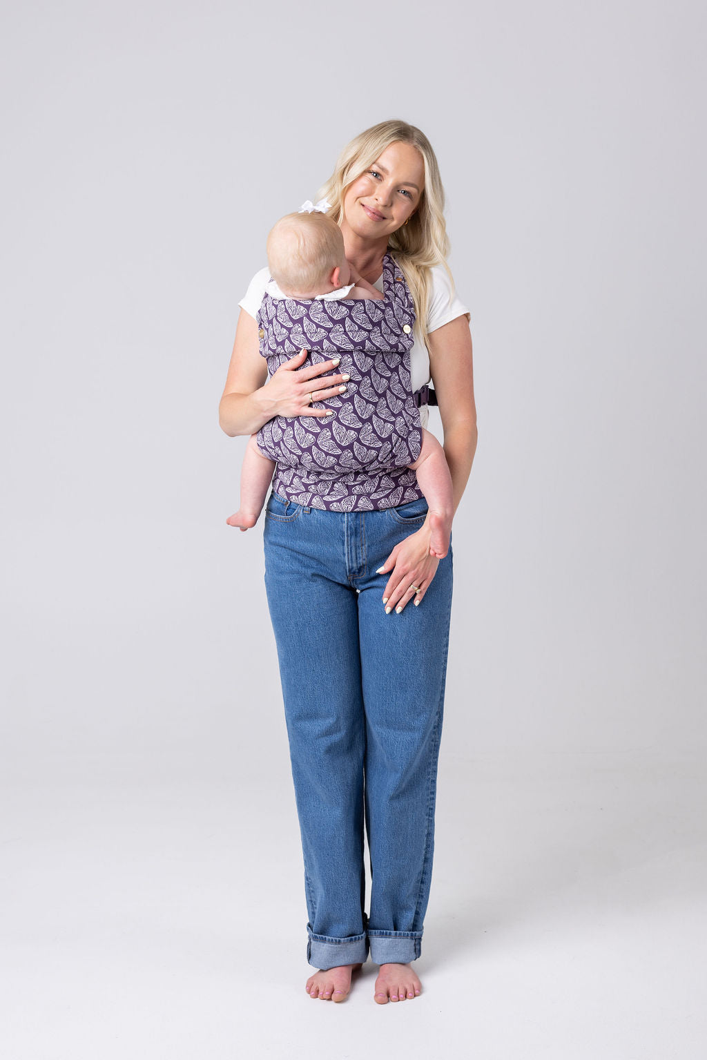 A light-skinned woman with long blonde hair, a white t-shirt, and medium-wash jeans carries a light-skinned, light-haired baby in the Mabē Monarch Buckle carrier in Violet Wings, which is dark purple with lighter butterfly wings patterned on it. She faces the viewer square on and her left hand rests flat on her left thigh and hip. She is barefoot.