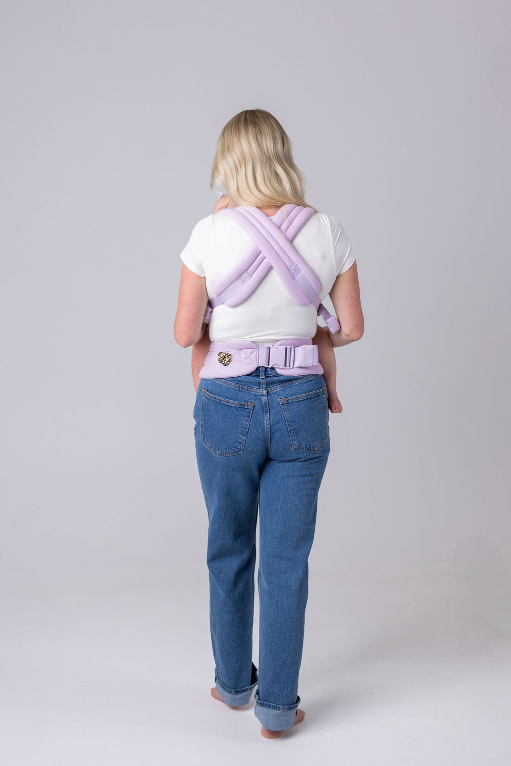 A person with an unseen face and long blonde hair faces away from the viewer. They have the crossed straps of the Mabē Monarch Buckle carrier in Lilac Skies across their back and the waistband buckled around their waist. They wear a white t-shirt and medium wash jeans. They are barefoot.