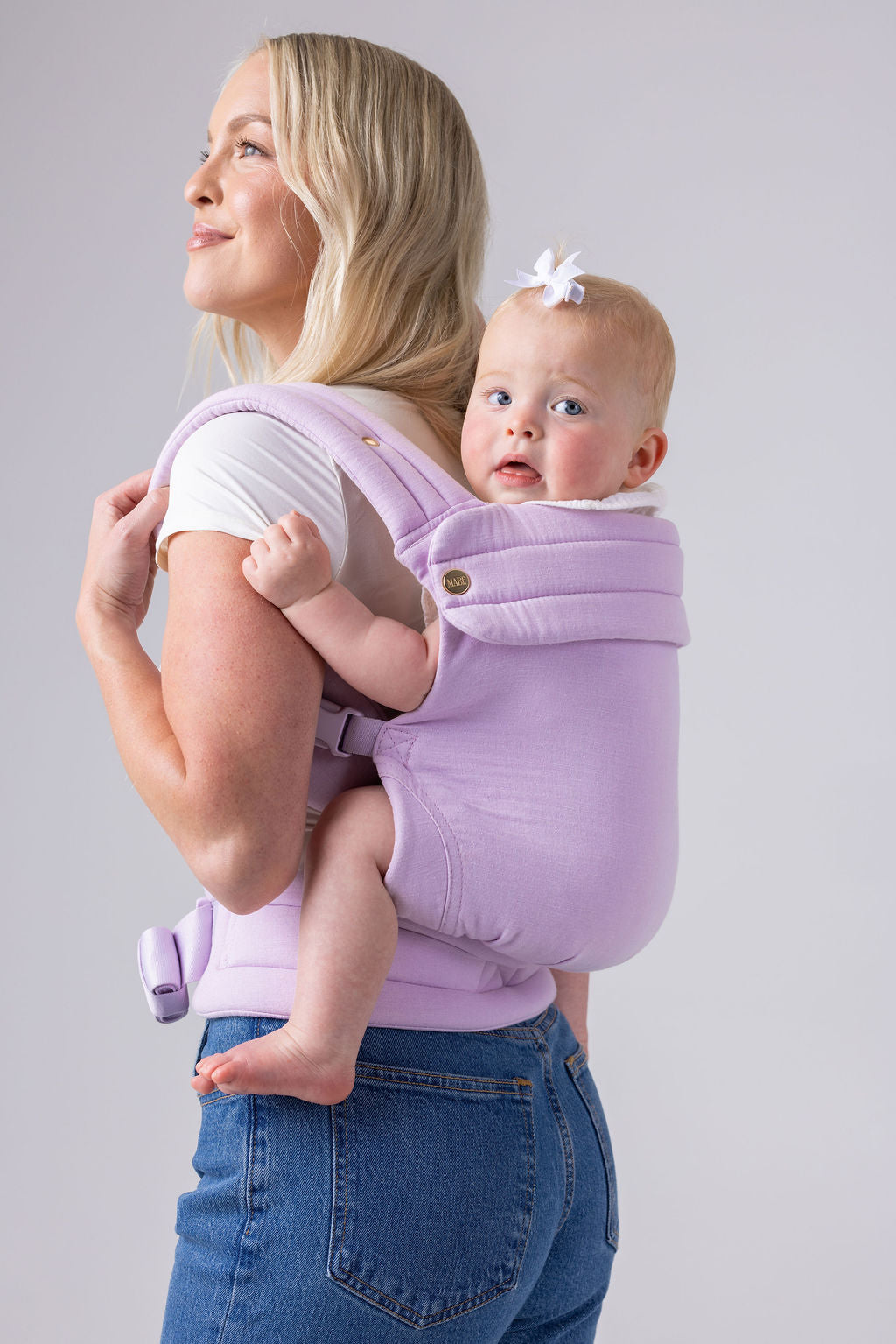 A photo of a light-skinned woman with long blonde hair, a white t-shirt, and medium-wash jeans. She carries a light-skinned, light-haired baby with a small white bow on her back in the Mabē Monarch Buckle carrier in Lilac Skies, which is solid lilac. She faces the left of the frame. The background is white or light gray.