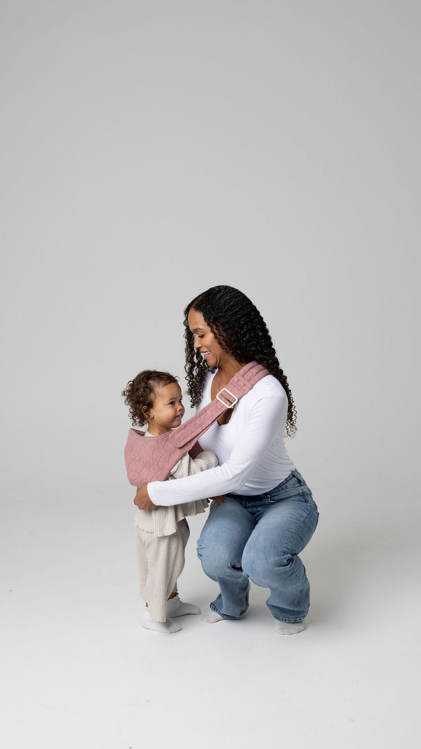 A tan-skinned woman with long, curly black hair kneels on the floor in a simple studio setting, gently supporting a toddler who stands close to her side, about to put the toddler up in a pink Mabē toddler sling with patterned butterfly wings. She wears a white long‑sleeve shirt and blue jeans, while the child is dressed in light, soft-toned clothing. The quilted pink sling crosses the woman’s shoulder and wraps around the child.