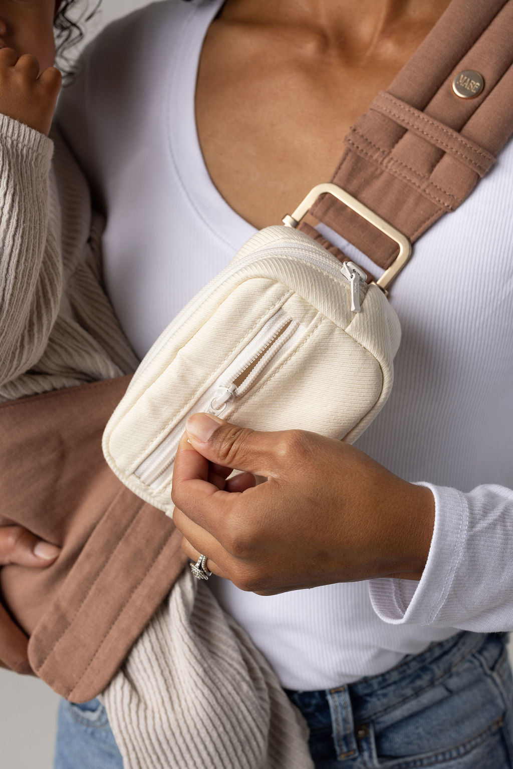 A cropped photo of a tan-skinned body wearing light-wash jeans and a white t-shirt with a mocha-colored Mabē Monarch toddler sling with a solid ivory accessory bag. A toddler is partly visible wearing cream colored co-ords.