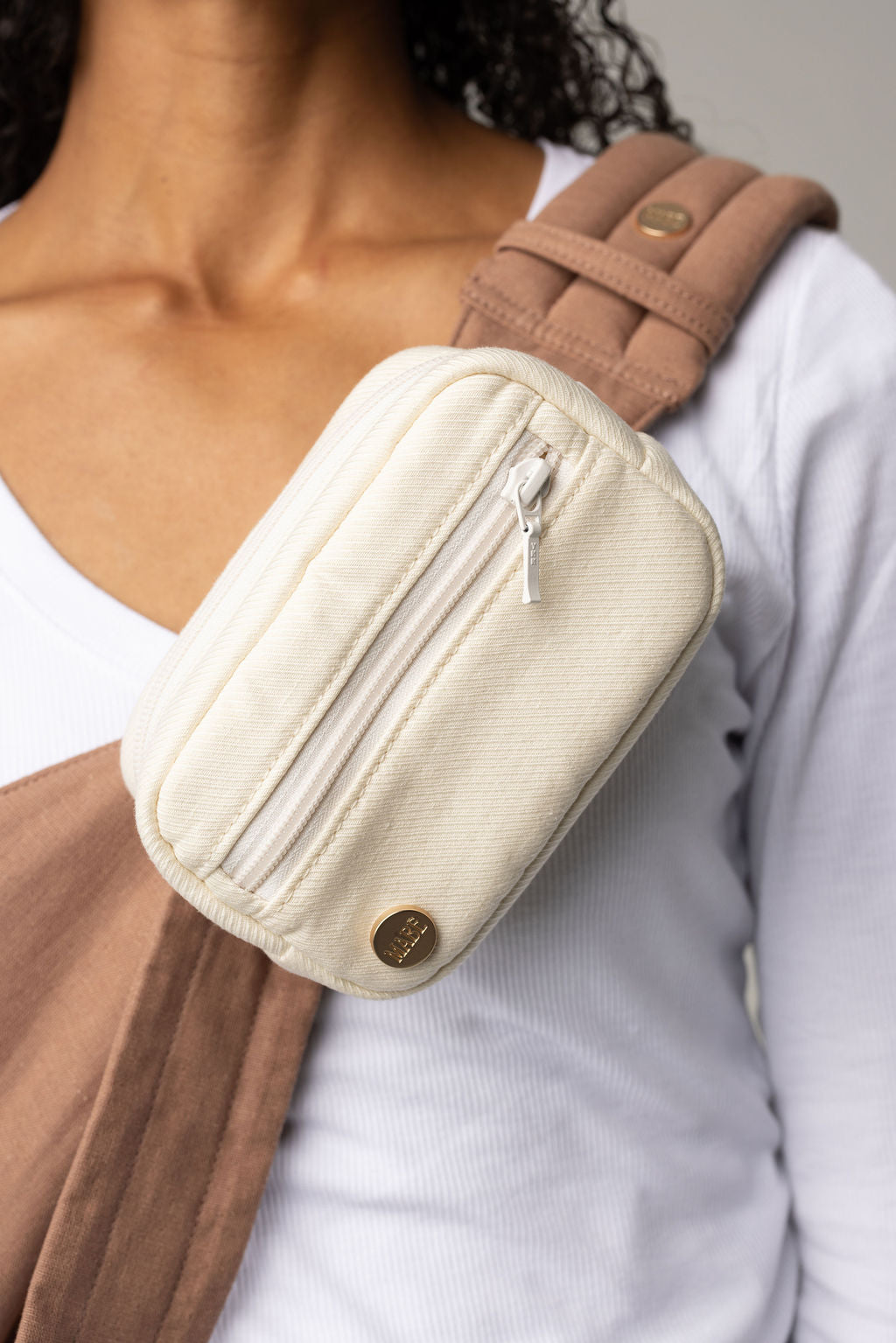 A cropped photo of a body wearing light-wash jeans and a white t-shirt with a mocha-colored Mabē Monarch toddler sling with a solid ivory accessory bag.