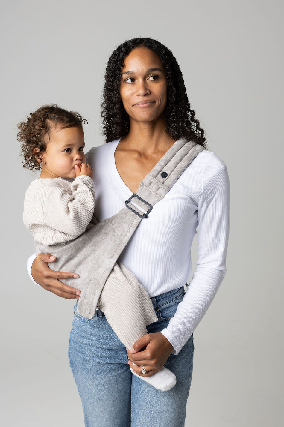 A brown-skinned woman with black curly long hair standing face-forward against a white background looking at her toddler in a sling carrier. She wears light-wash jeans and a white t-shirt and has a toddler  in a light gray Ashland Camo Mabē Monarch toddler sling. It has a padded, quilted strap and a seat for the child to sit in, and a gold ladder-lock adjuster. The toddler is wearing coordinating cream sweater and pants and white socks and is looking to the right of the frame.