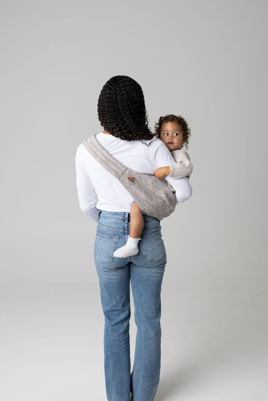A brown-skinned woman with curly black hair shown full height facing away from the viewer is holding a toddler with curly brown hair in a light gray Ashland Camo Mabē toddler sling. The woman wears a long-sleeved white shirt and mid-wash jeans, and the toddler wears cream-colored knit coordinates and white lettuce-trim socks.