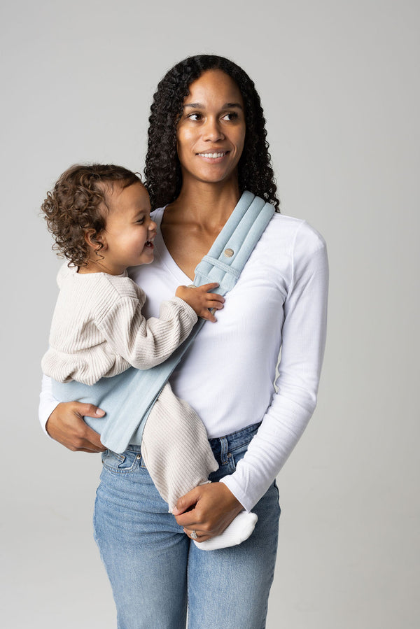 A brown-skinned woman with long, black, curly hair holding a tan-skinned toddler with brown, curly hair in a light blue Dream Wash Denim Mabē toddler sling. The woman wears a long-sleeved white shirt and mid-wash jeans, and the toddler wears cream-colored knitted coordinating long-sleeved top and bottom and white socks. They stand against a white background.