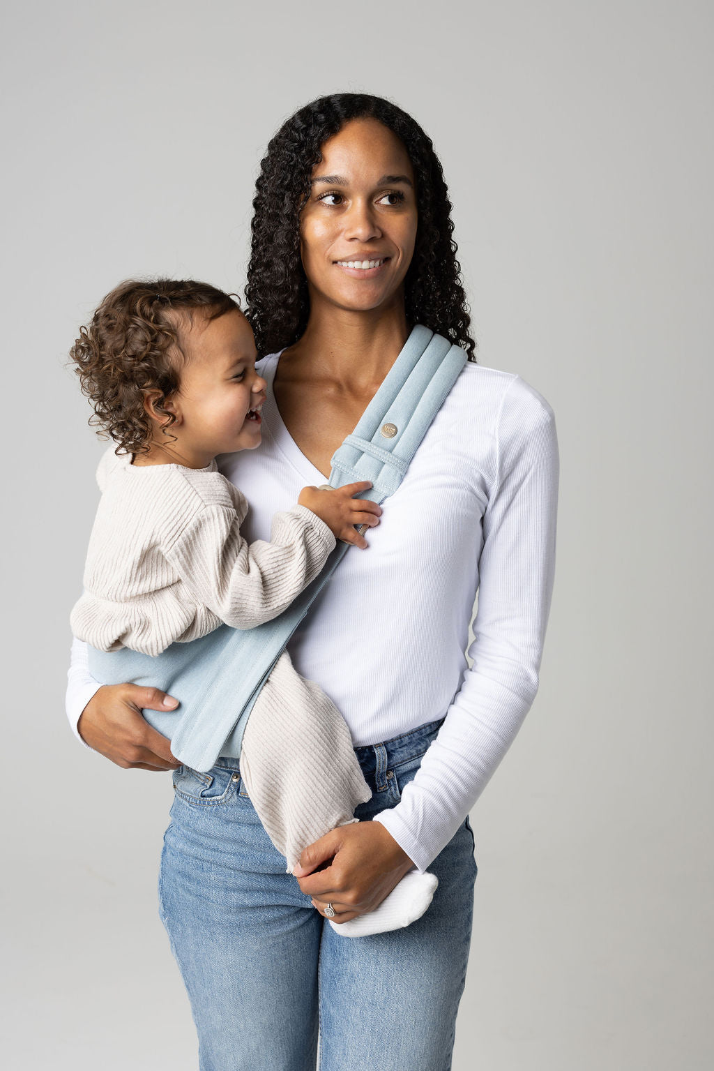 A brown-skinned woman with long, black, curly hair holding a tan-skinned toddler with brown, curly hair in a light blue Dream Wash Denim Mabē toddler sling. The woman wears a long-sleeved white shirt and mid-wash jeans, and the toddler wears cream-colored knitted coordinating long-sleeved top and bottom and white socks. They stand against a white background.