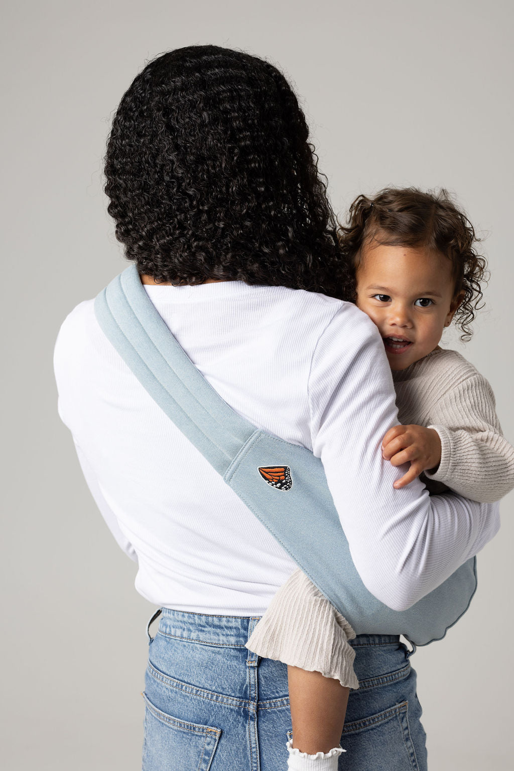 A brown-skinned woman facing away from the viewer. She has long, black, curly hair and is holding a tan-skinned toddler with brown, curly hair in a light blue Mabē Dream Wash Denim toddler sling. The woman wears a long-sleeved white shirt and mid-wash jeans, and the toddler wears cream-colored knitted coordinating long-sleeved top and bottom and white socks. They stand against a white background.