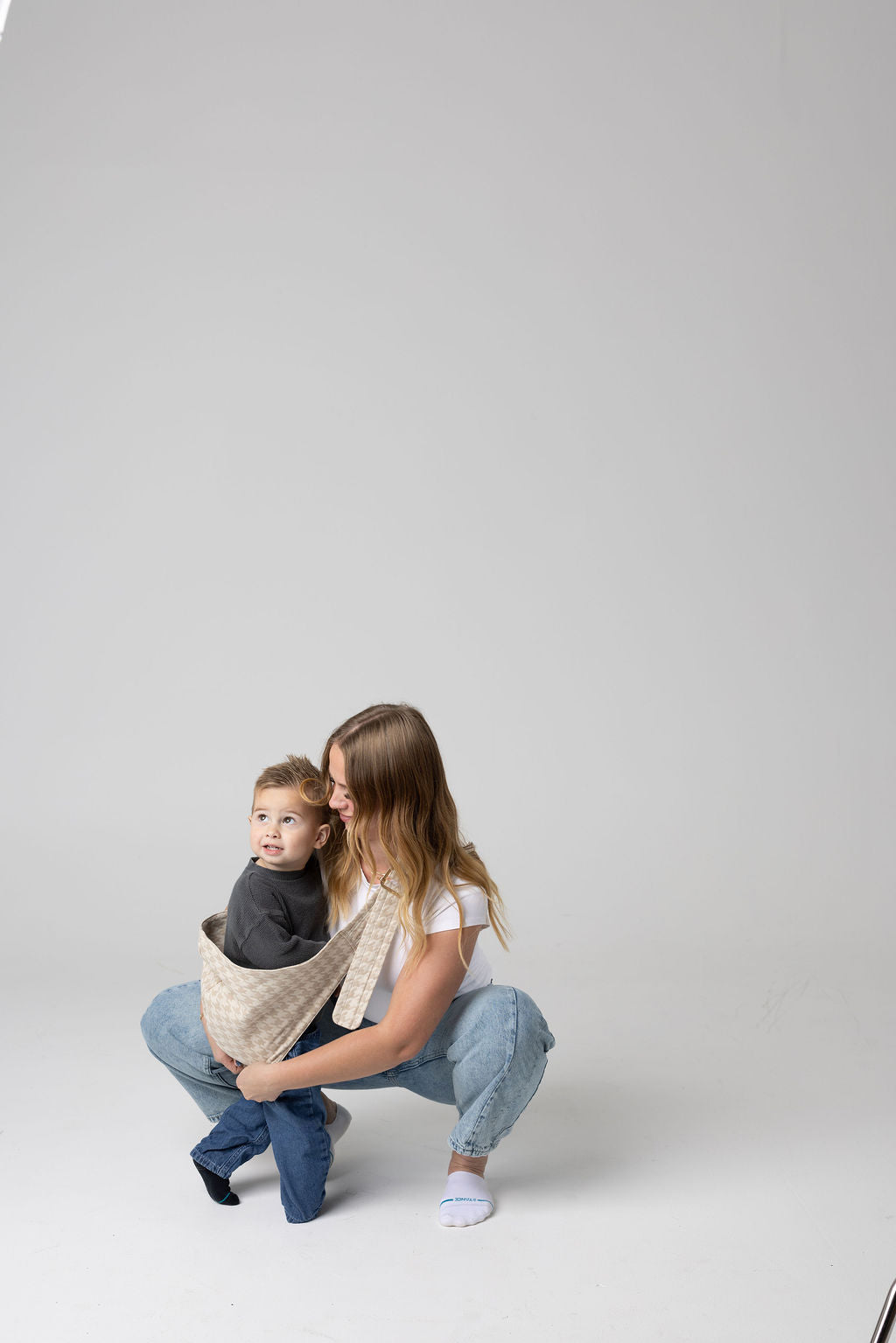 A brown-haired woman with blonde highlights crouched against a white background. She wears light-wash jeans and a white t-shirt and looks at a toddler boy being scooped uin the  Mabē Monarch houndstooth-patterned toddler sling in beige and cream. It has a padded, quilted strap. The toddler is wearing a gray sweater and medium-wash jeans and black and gray socks and is looking to the upper-left of the frame.